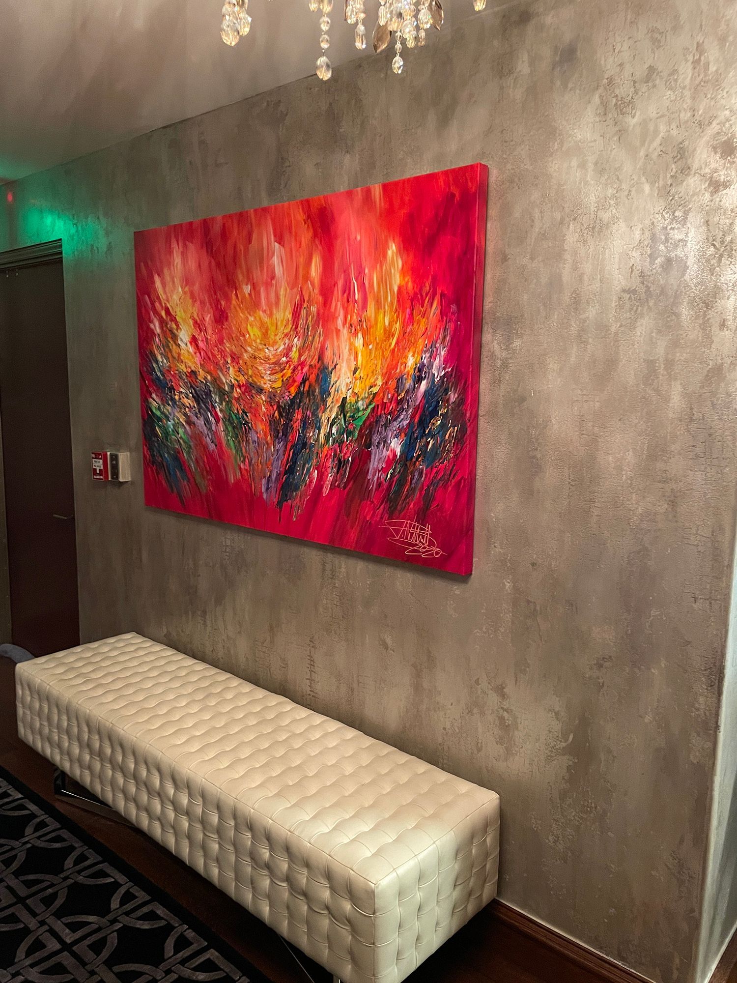 Abstract red and yellow painting above a white, quilted bench. Dark rug on the floor, on a textured gray wall.
