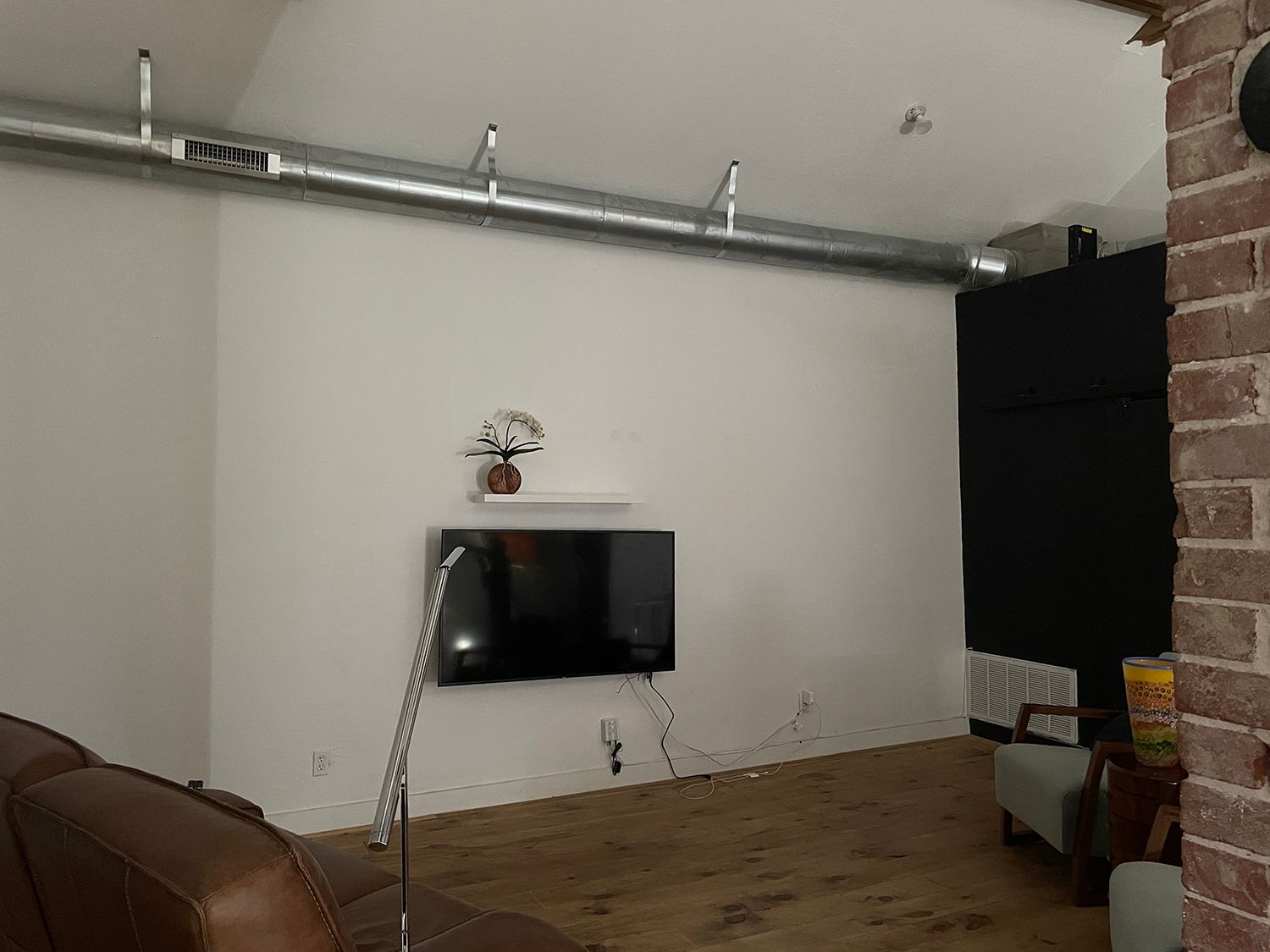 Living room with TV mounted on white wall, exposed ductwork, brick pillar, and wood floor.
