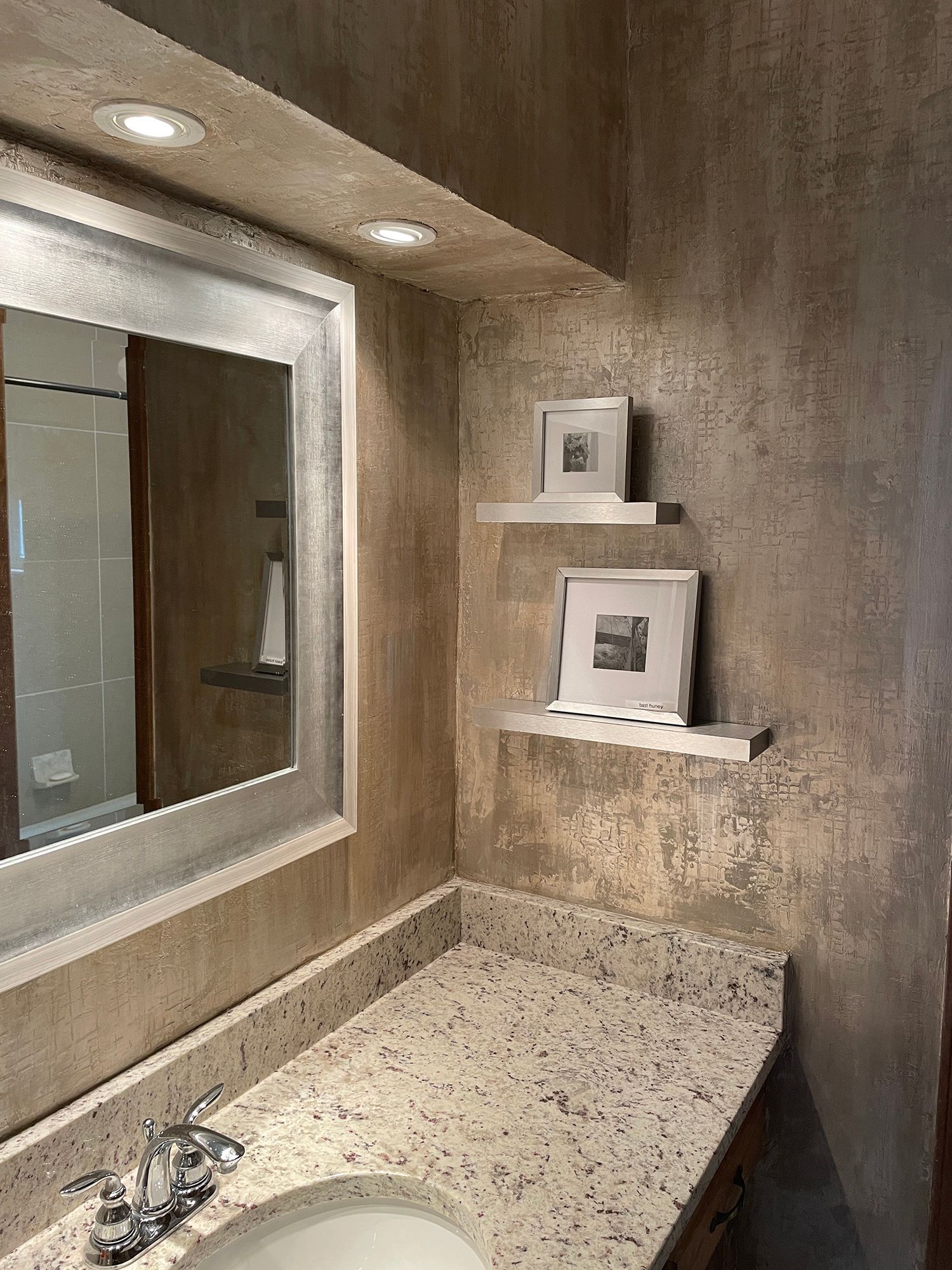 Bathroom corner with textured beige walls, granite countertop, mirror, and two floating shelves with framed artwork.