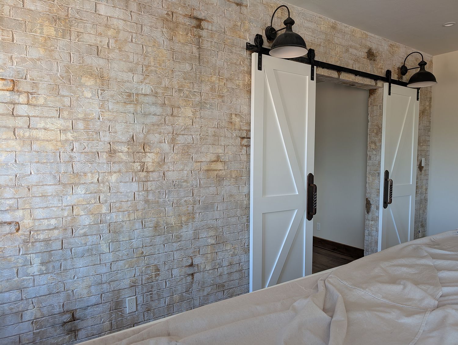 White barn doors on a brick wall, black track, light fixtures. A white bed is in the foreground.