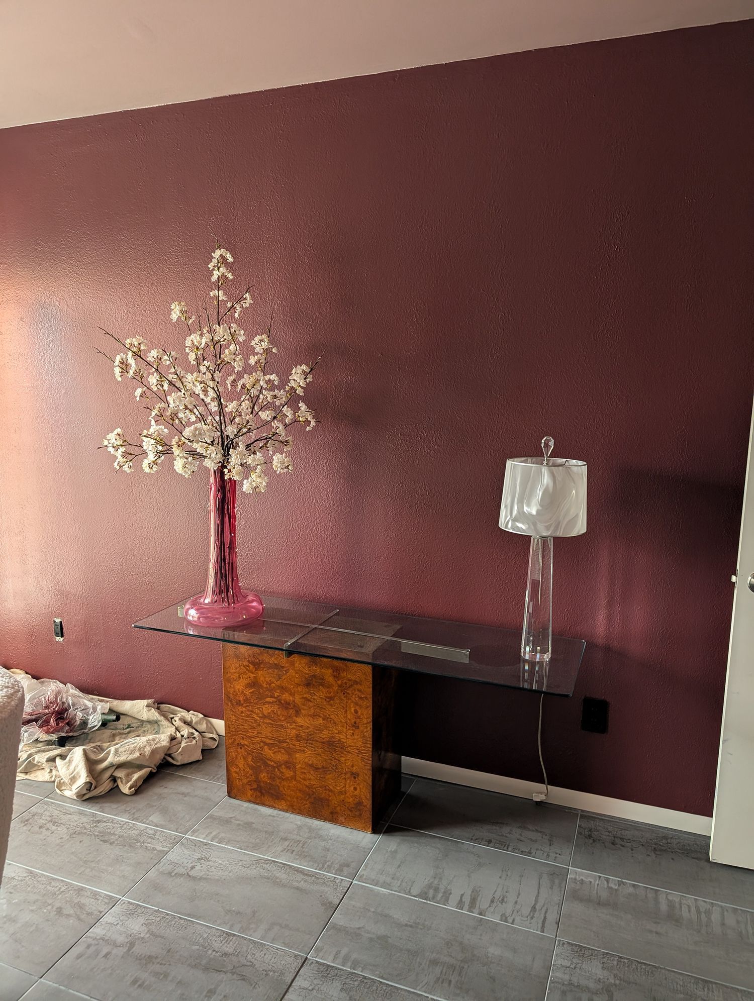 Desk with a brown base and glass top, a cherry blossom arrangement in a pink vase, and a lamp against a mauve wall.