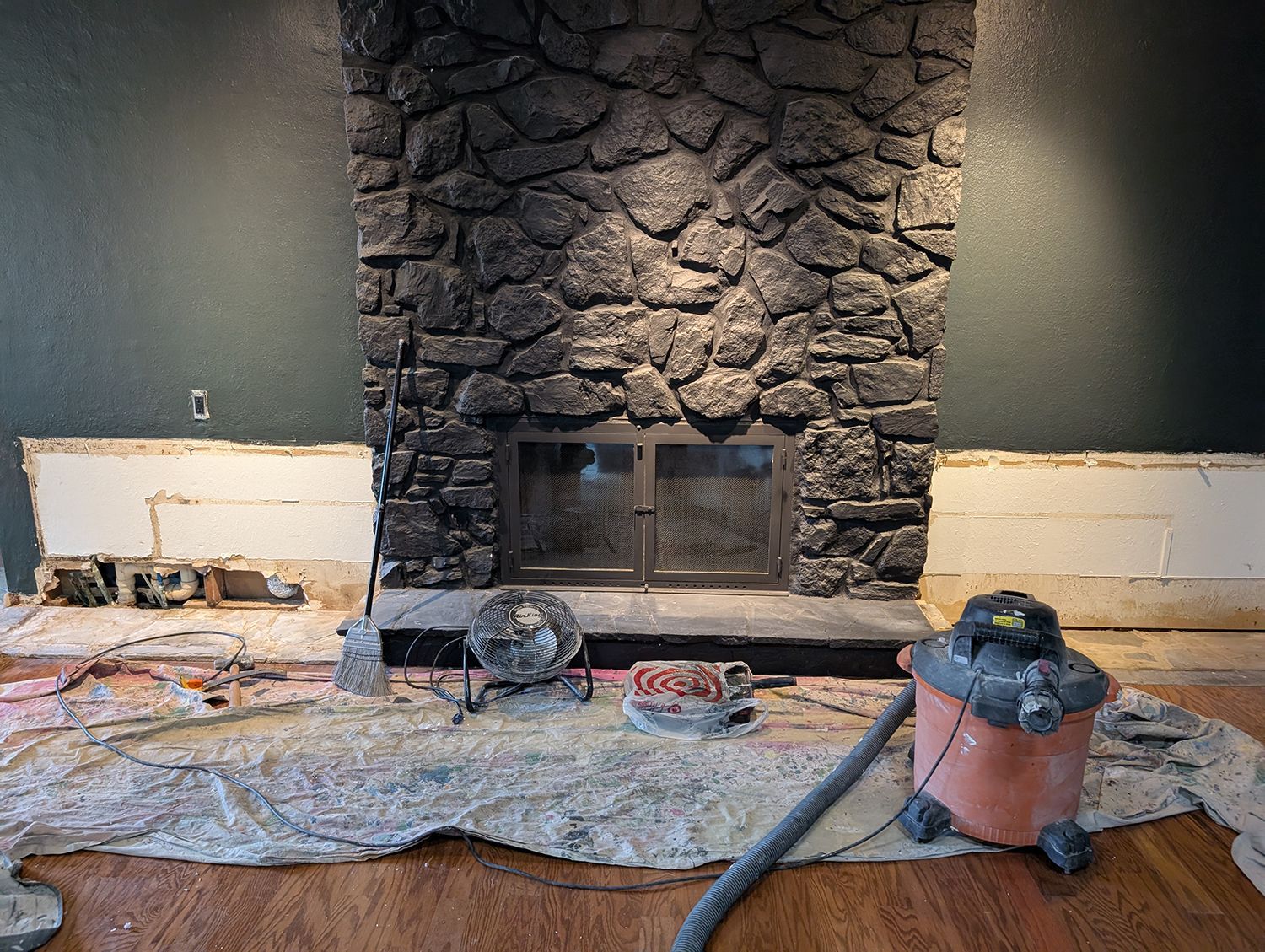 Fireplace being renovated; stone surround, dark metal firebox. Tools and debris on floor.