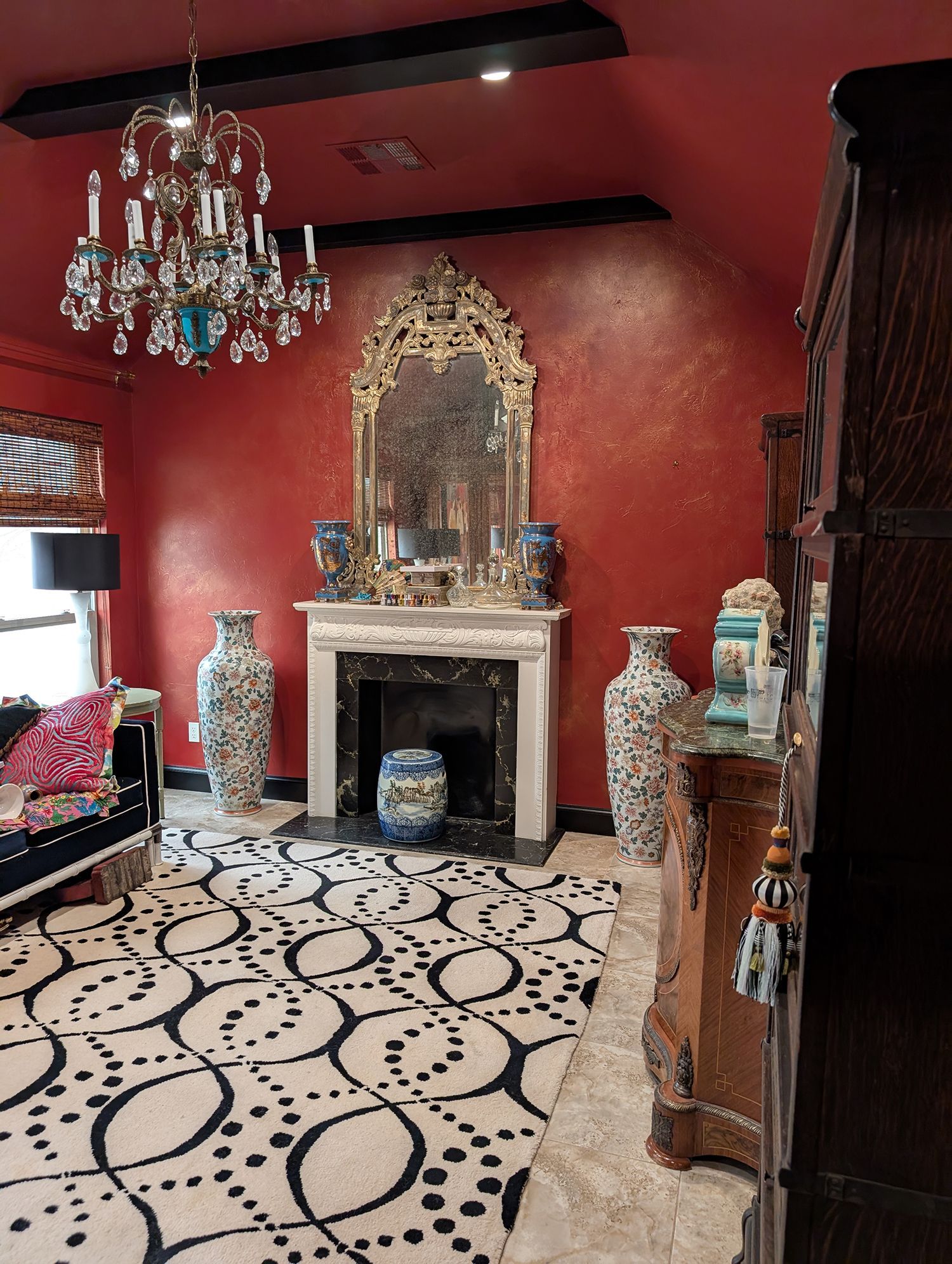 Room with ornate decor: red walls, chandelier, fireplace with gold mirror, patterned rug, tall vases.