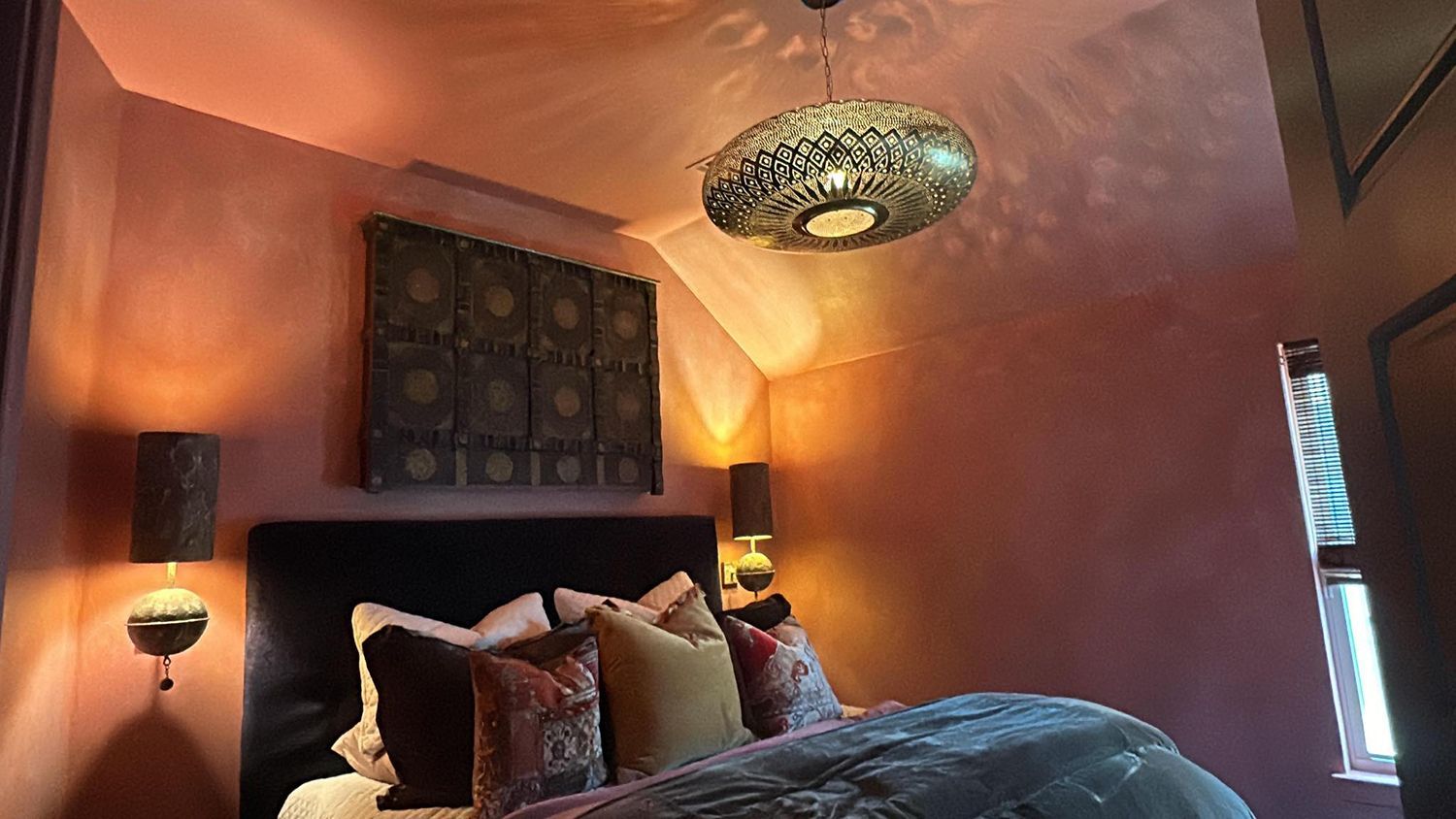 Cozy bedroom with pink walls, ornate lamps, and a decorative pendant light over a bed with pillows.