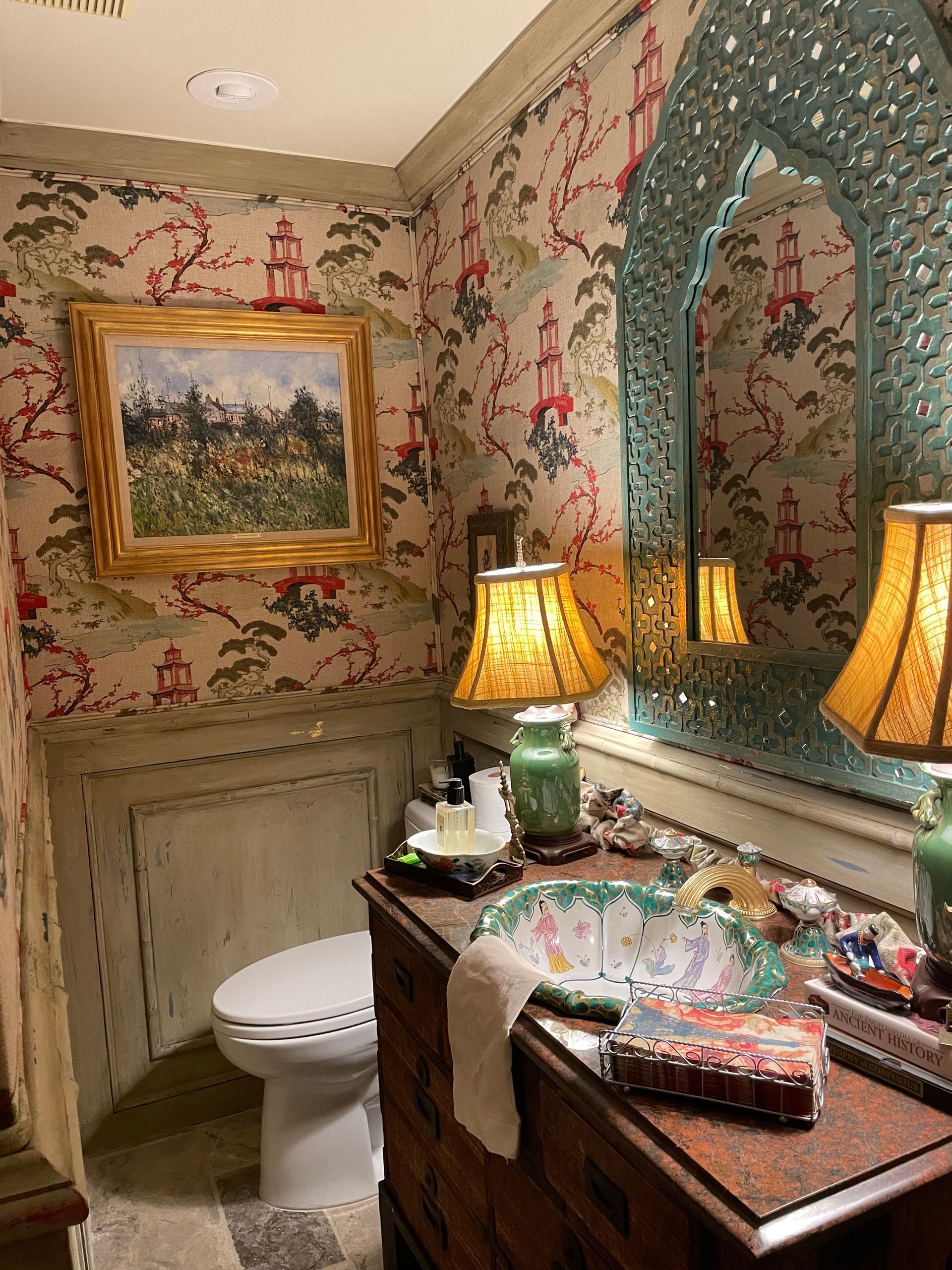 A decorated powder room. Floral wallpaper, ornate mirror, and antique vanity with a landscape painting.