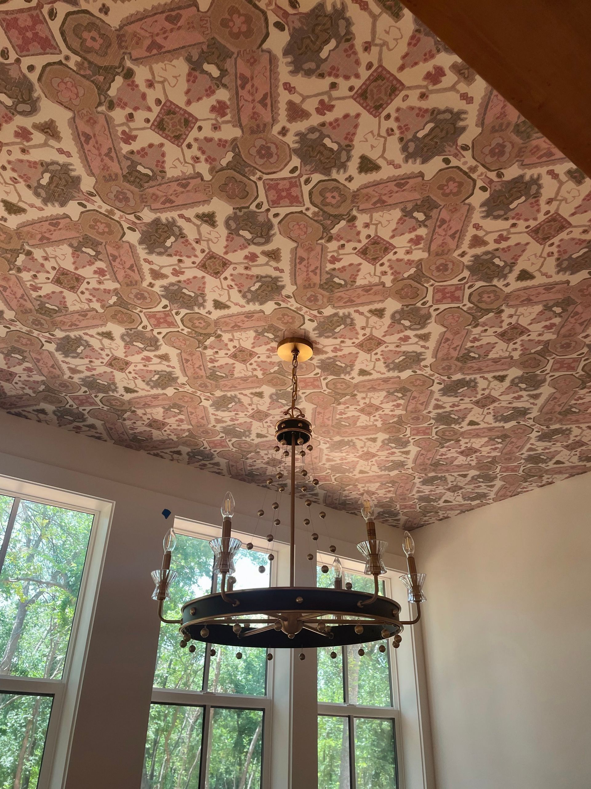Chandelier hangs from patterned ceiling, with light and dark pink, and gray designs. Windows and trees in background.