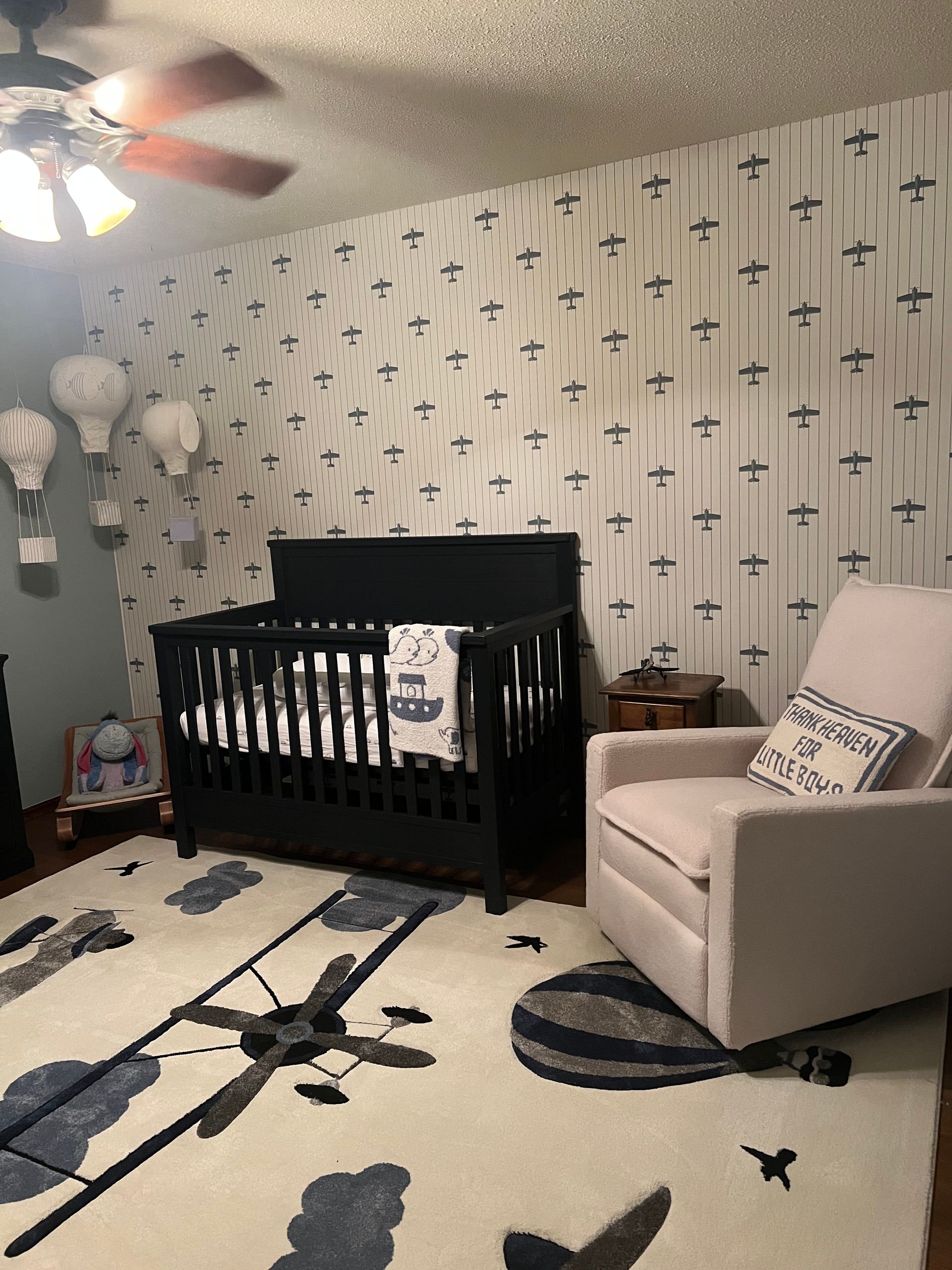 Nursery with airplane-themed decor: black crib, beige rocking chair, airplane rug, wallpaper with airplanes.