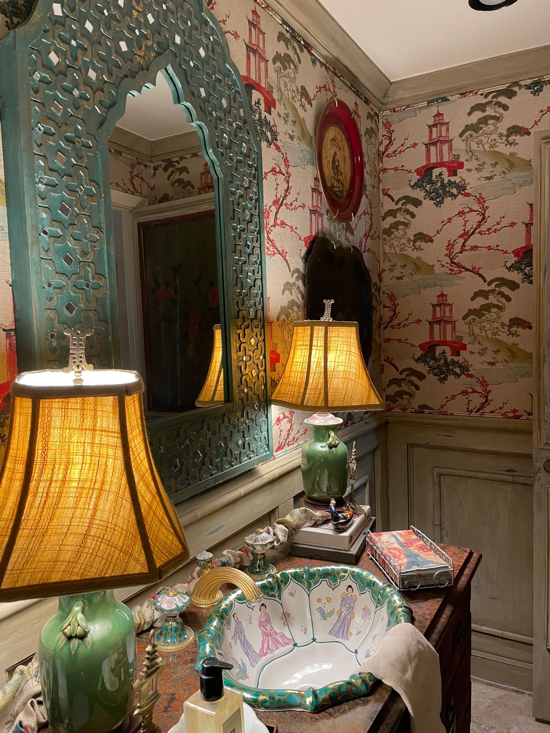 Vanity with ornate turquoise mirror, floral sink, wallpaper, and lamps.