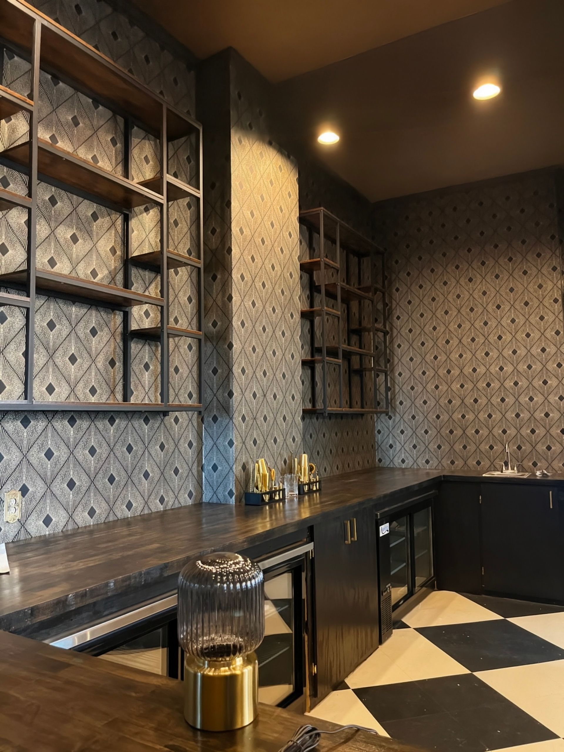Bar area with dark wood counters, shelves, and patterned wallpaper. Black and white checkered floor.