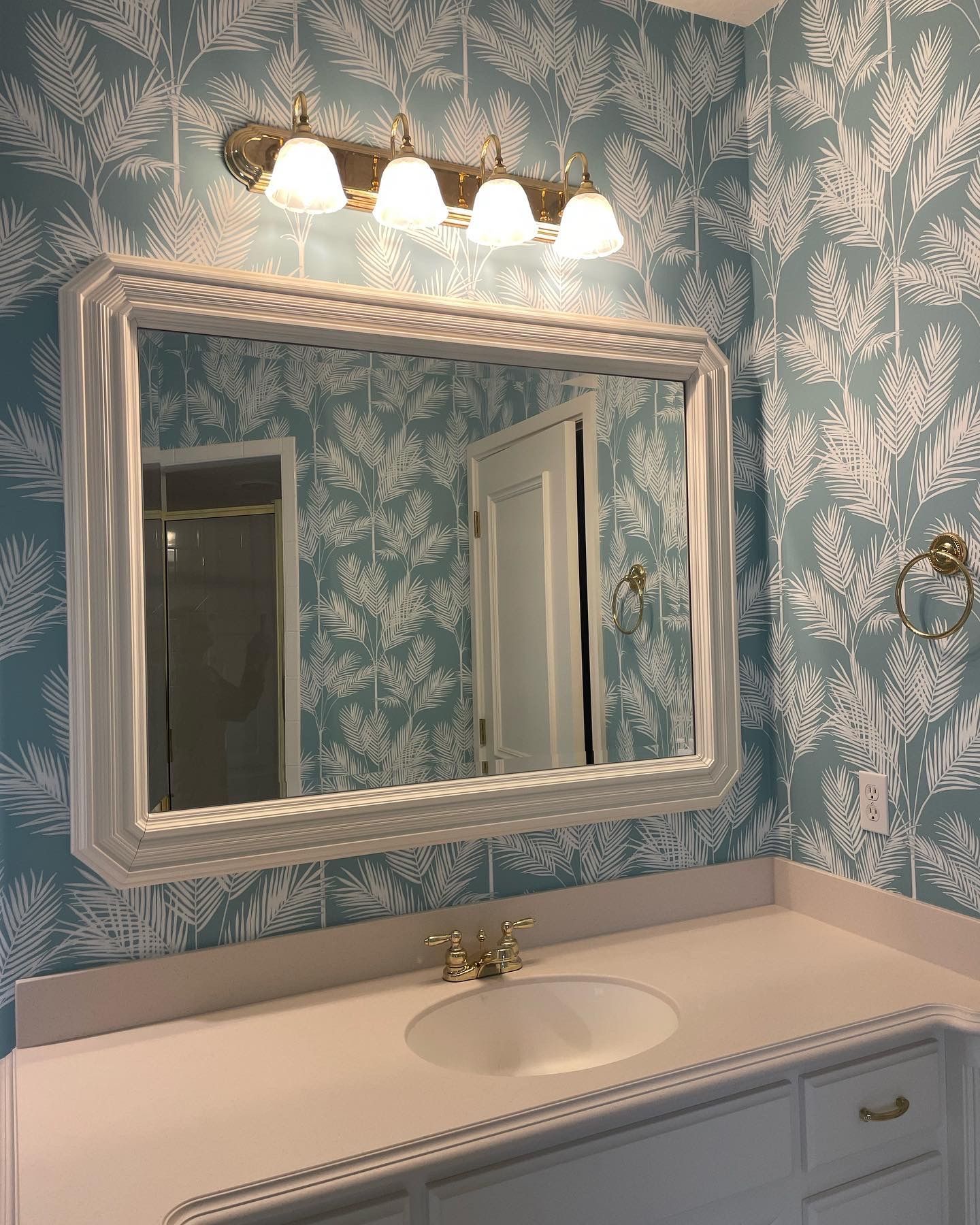 Bathroom with white countertop, oval sink, and floral wallpaper. Large mirror, light fixture, and gold hardware.