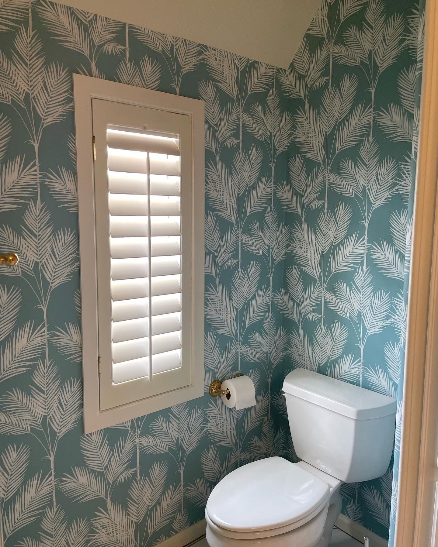 Small bathroom with turquoise and white wallpaper, white toilet, and window with shutters.