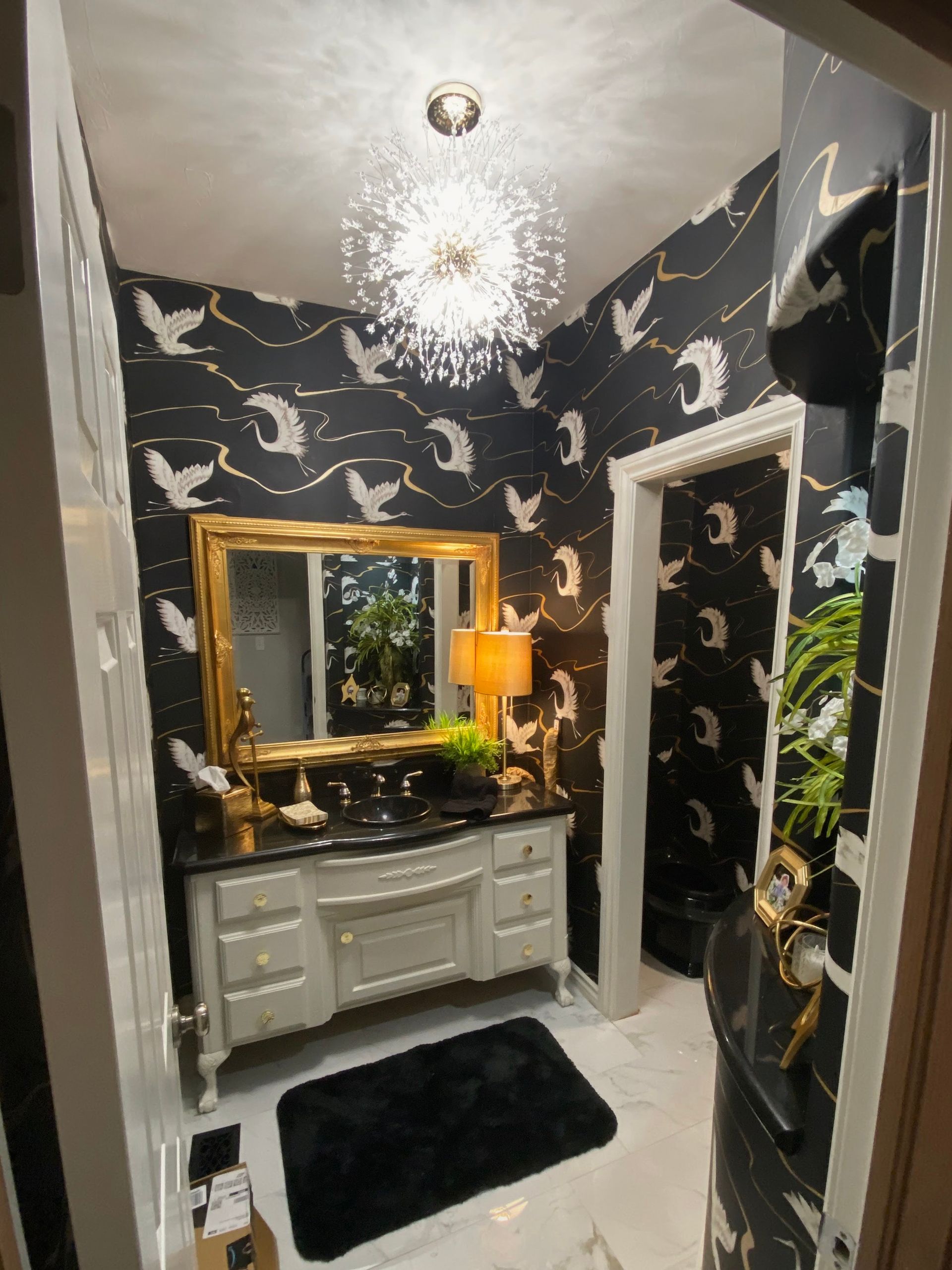 Small bathroom with swan-patterned wallpaper, ornate mirror, cream vanity, and sparkly chandelier.