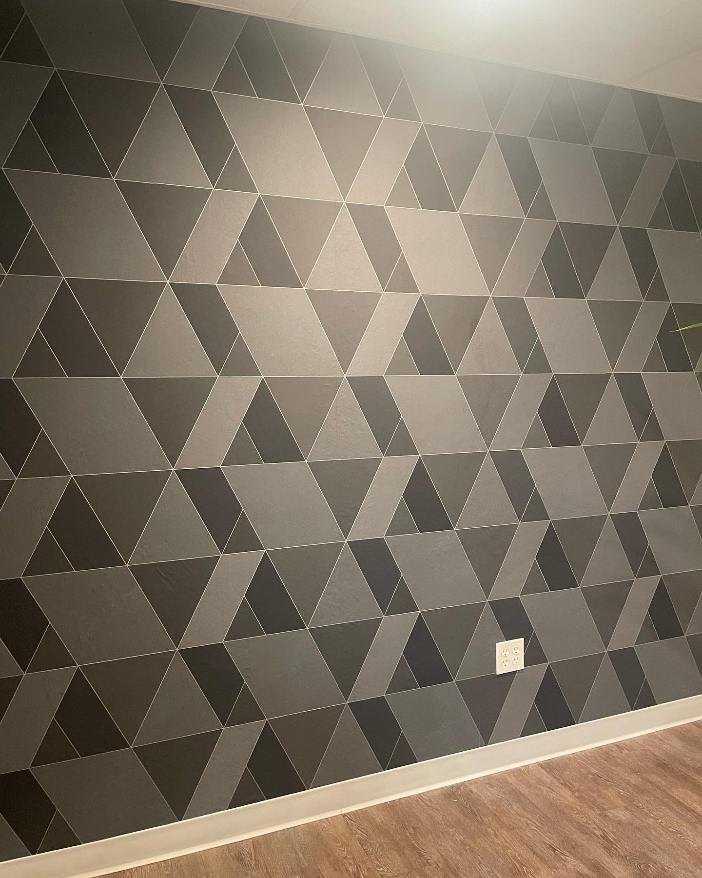 Wall with geometric 3D shapes in shades of gray, with a power outlet on the wall and wooden flooring.
