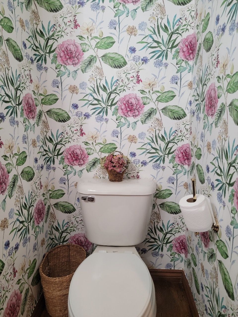 Toilet in a small room with floral wallpaper and a small decorative flower arrangement on the tank.
