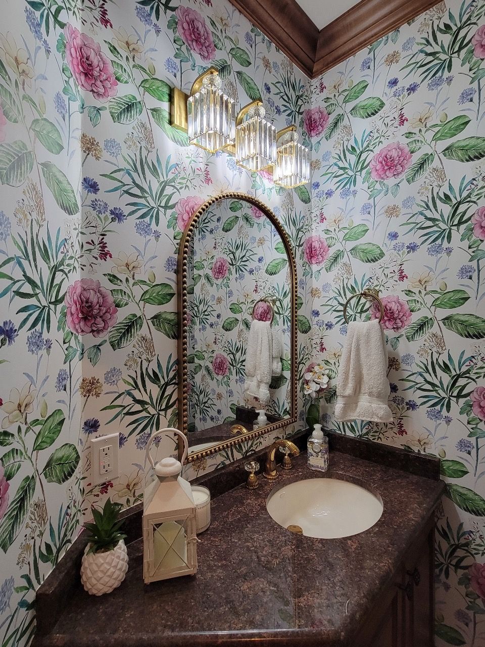 Floral wallpapered powder room with gold accents. Includes mirror, sink, and decorative items.
