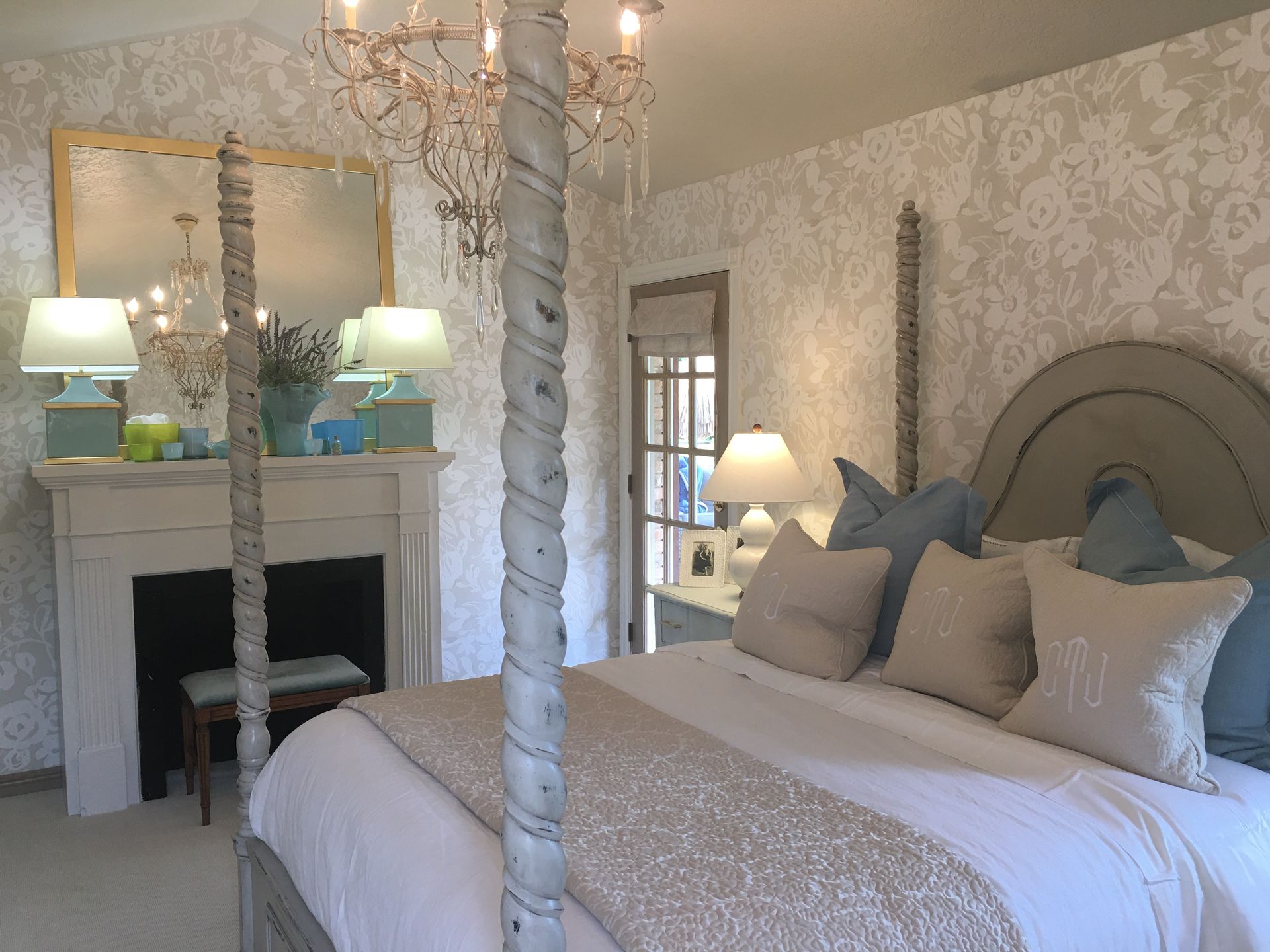 Elegant bedroom with bed, fireplace, chandelier, and patterned wallpaper. Soft tones of beige and blue.