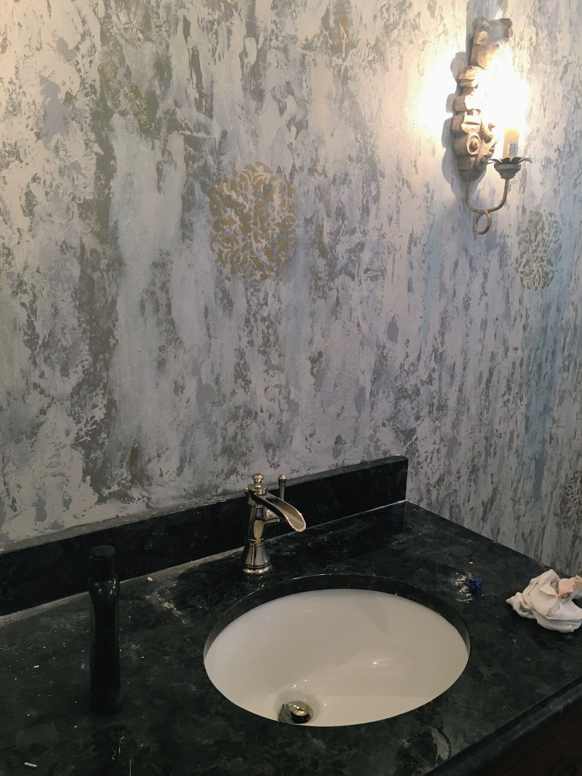 Vanity with dark countertop and sink. Textured, blue-gray wall with sconce.
