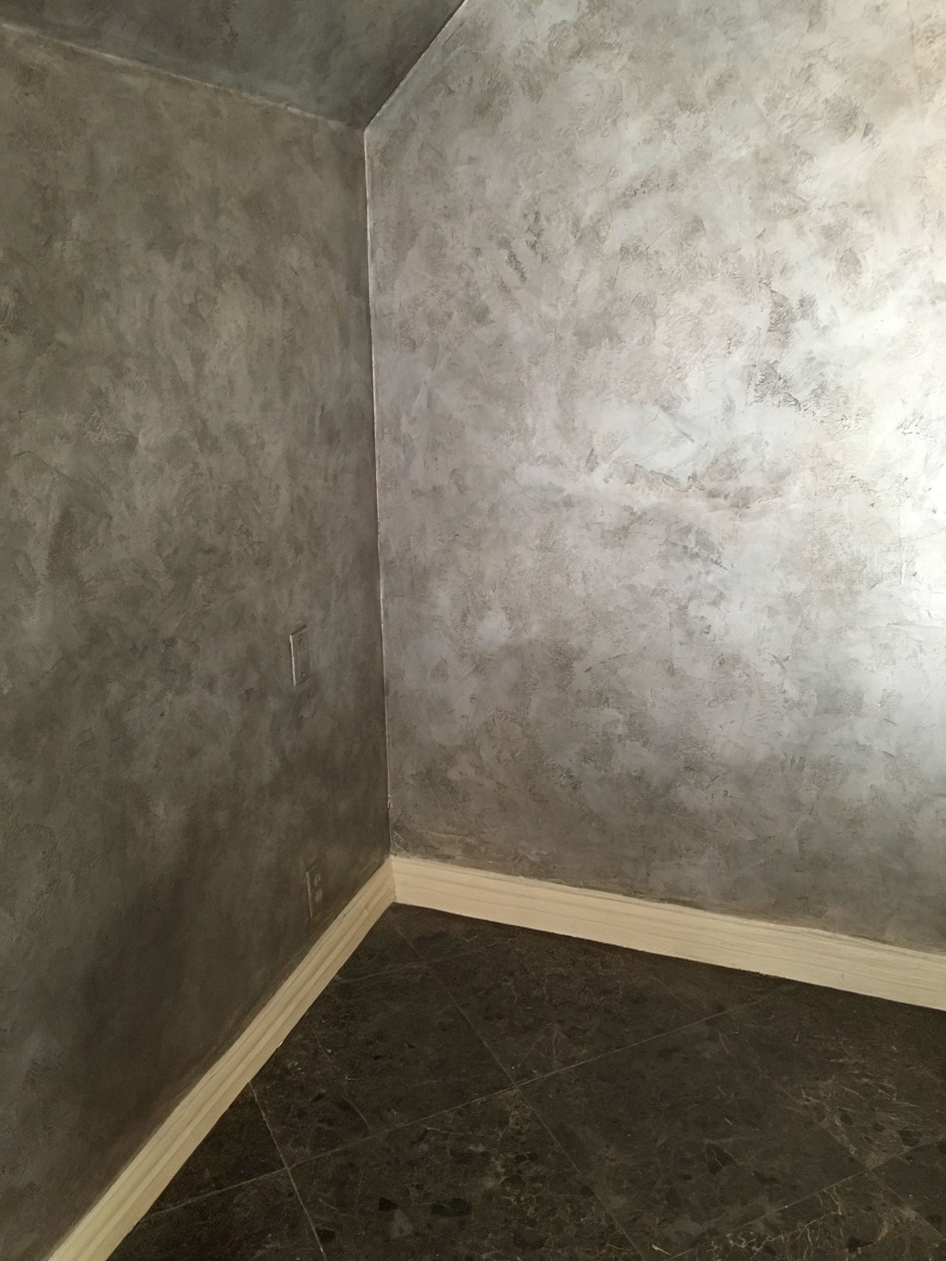 Corner of a room with textured gray walls and light-colored baseboards. Dark floor.