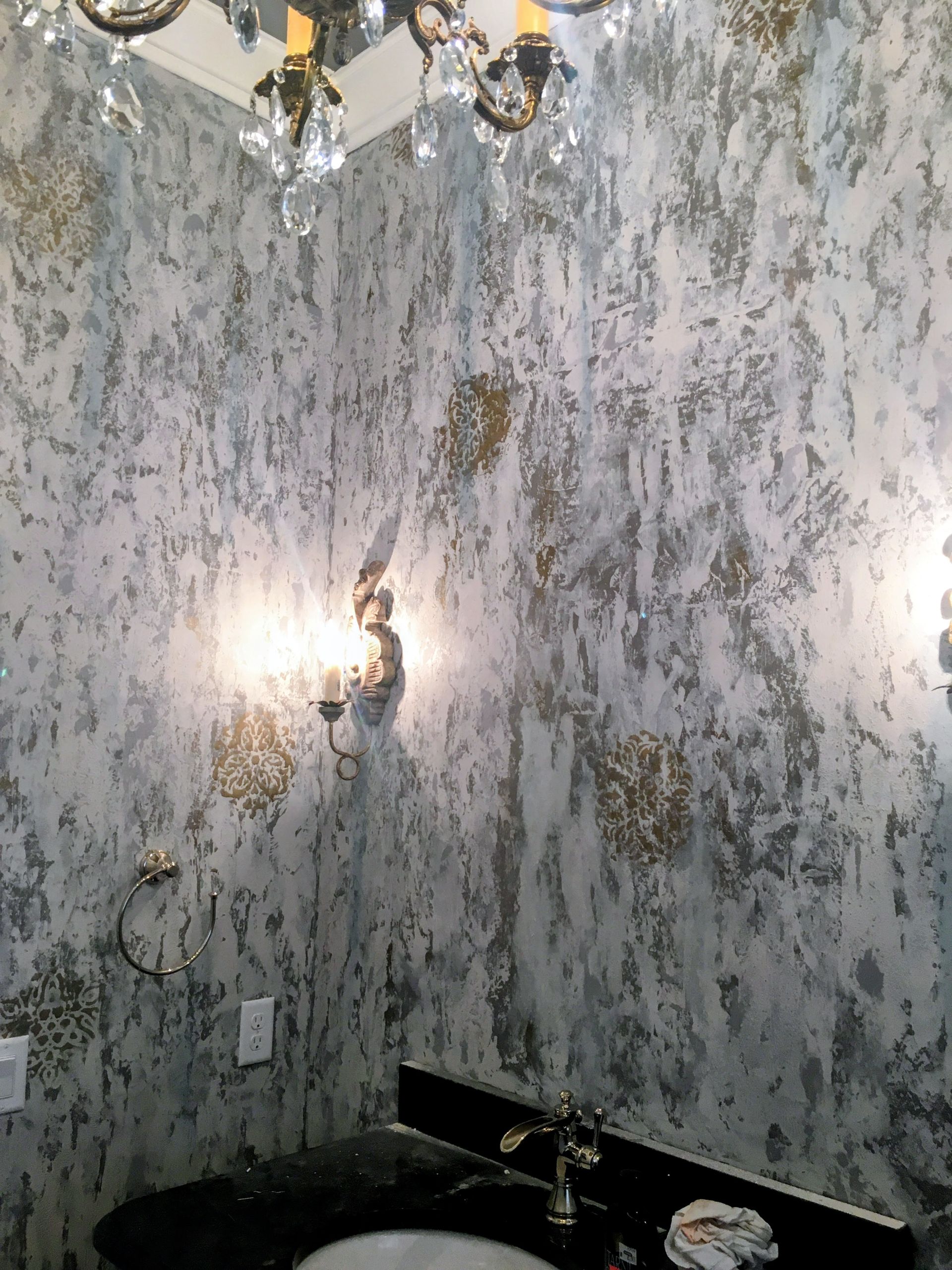 Bathroom with textured silver and gold wall finish, sconces, and a black countertop.