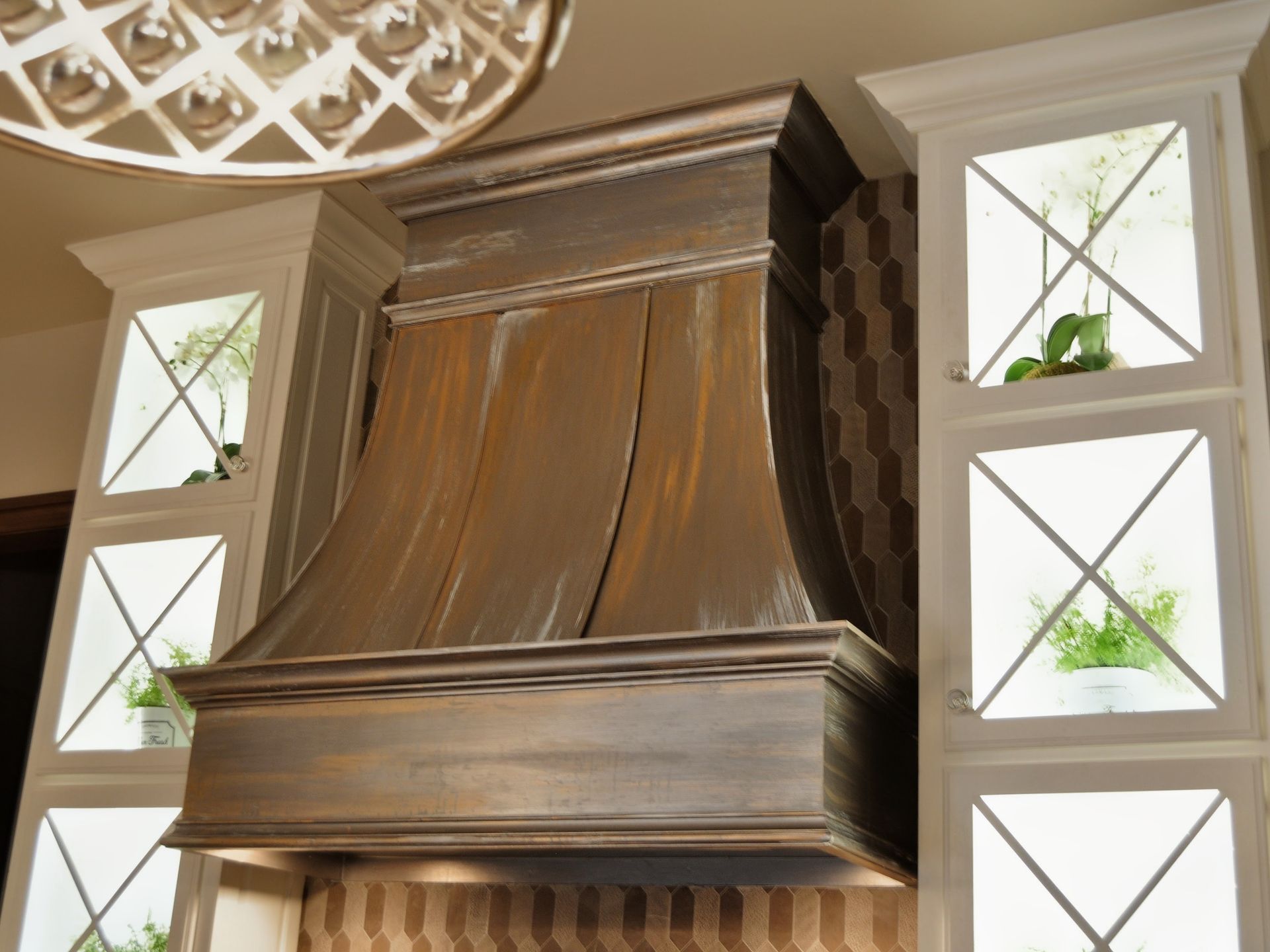 Range hood in a kitchen, with a weathered brown finish and flanked by white cabinets with glass inserts.