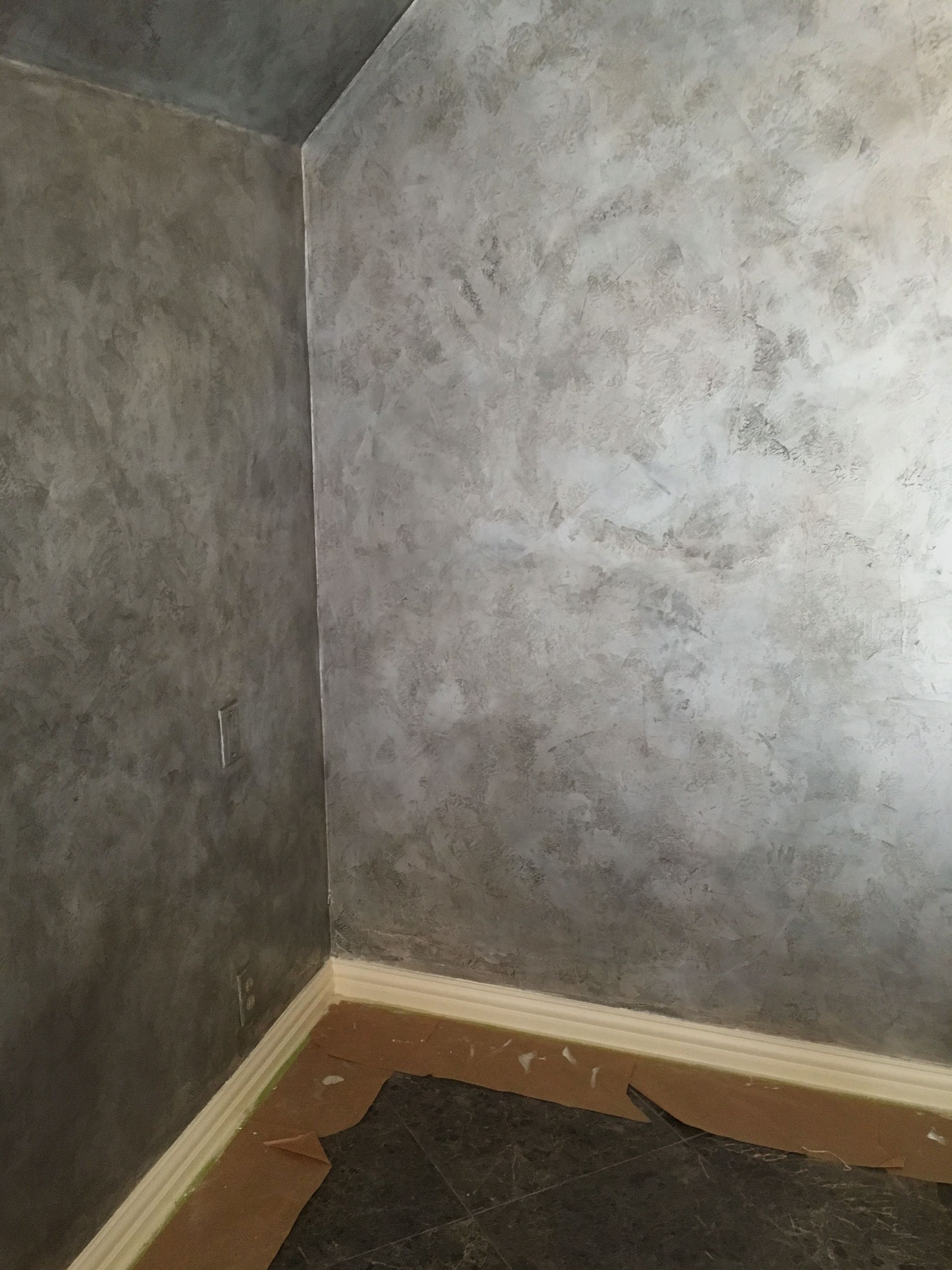 Corner of room with mottled gray-brown walls, light-colored baseboard, and a dark floor covering.