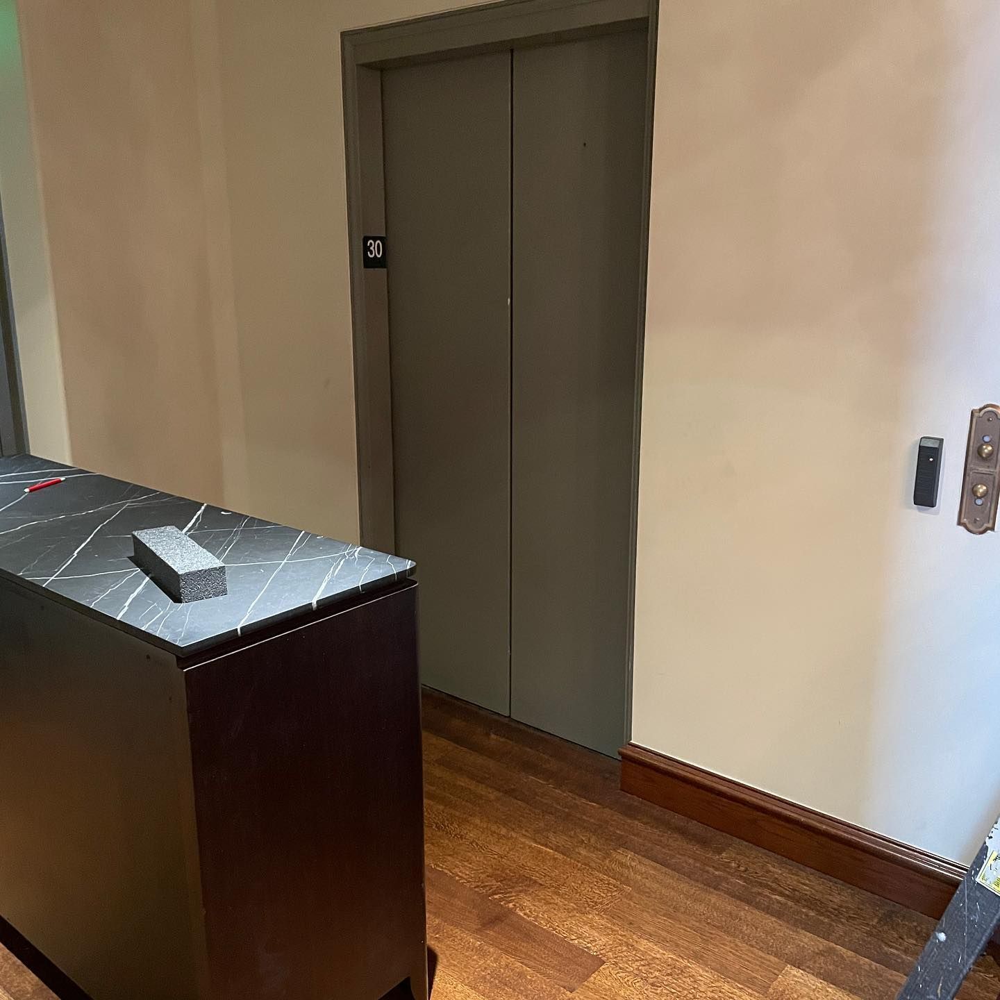 Elevator with closed green doors, wooden floor, brown cabinet, beige walls, and call button.