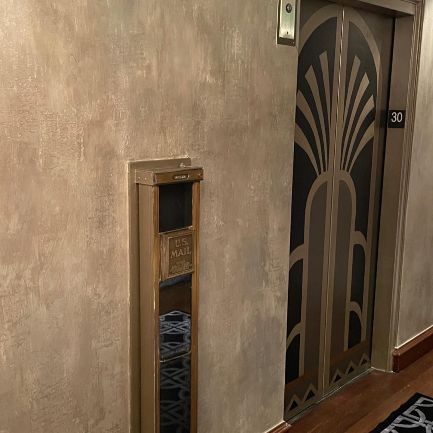 An art deco elevator door and mailbox in a hallway with textured tan walls and wooden floor.