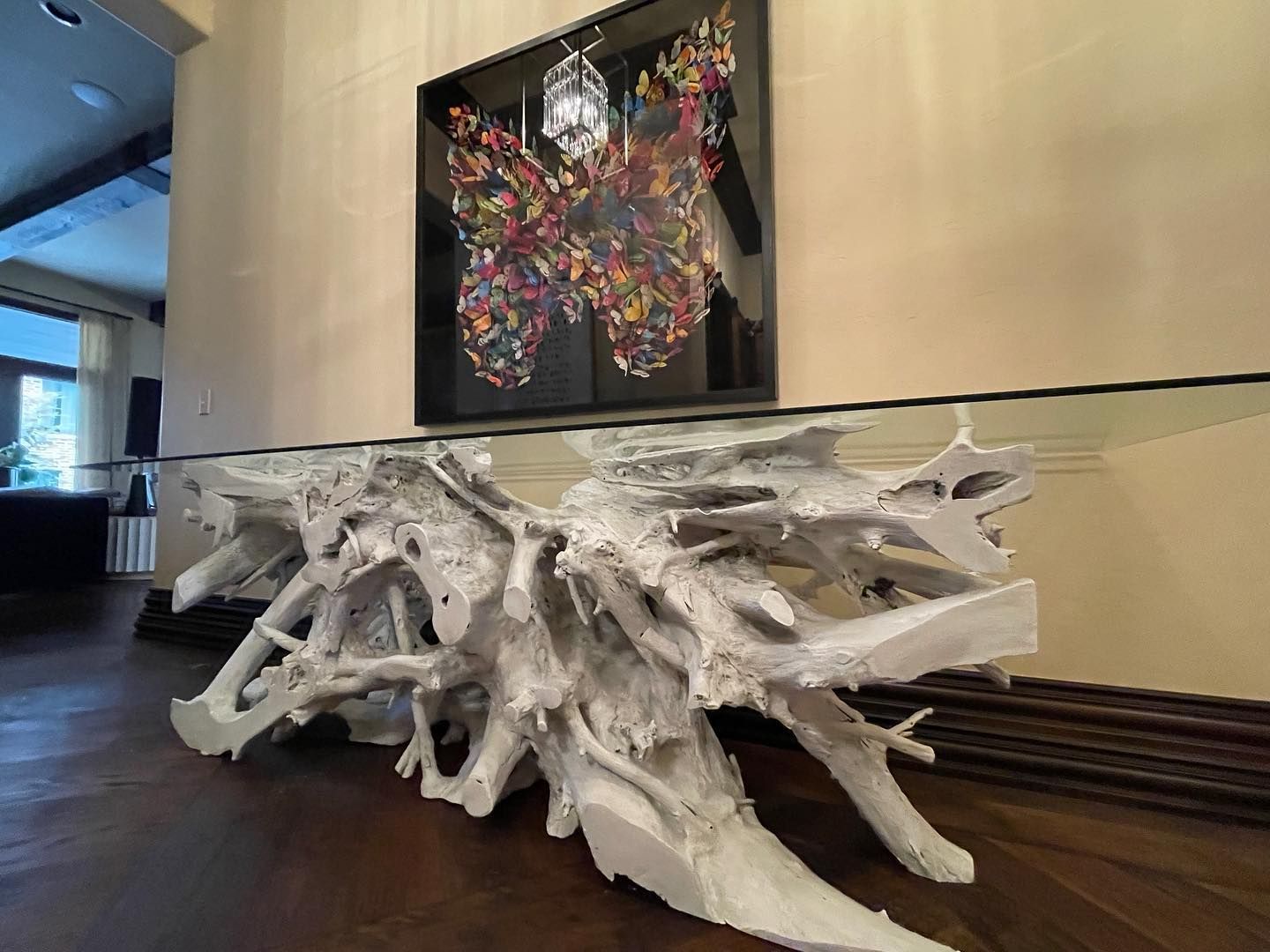 White root-like table supporting a clear glass top, beneath a framed abstract butterfly art piece.