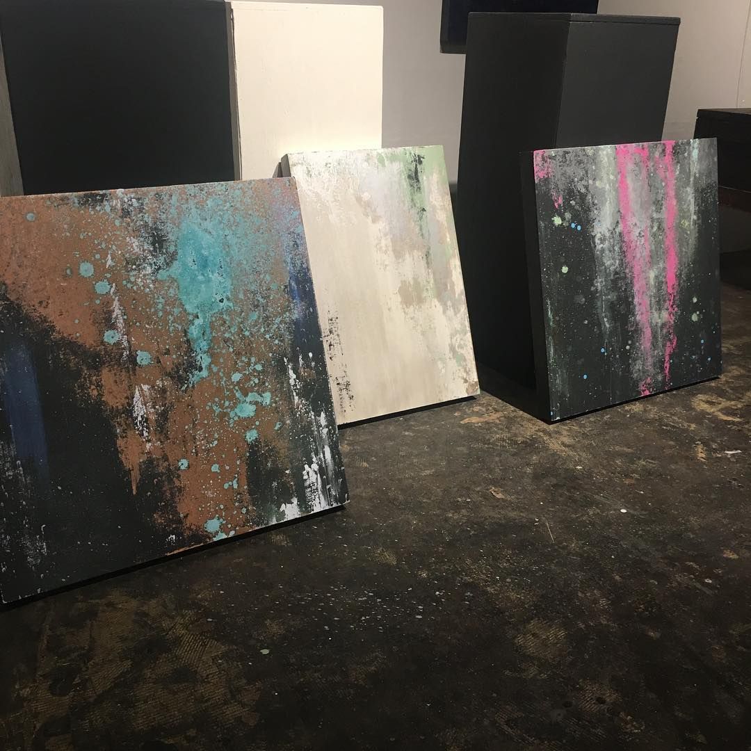 Four abstract paintings in a studio. Each canvas has splattered paint in various colors; the setting is dark.
