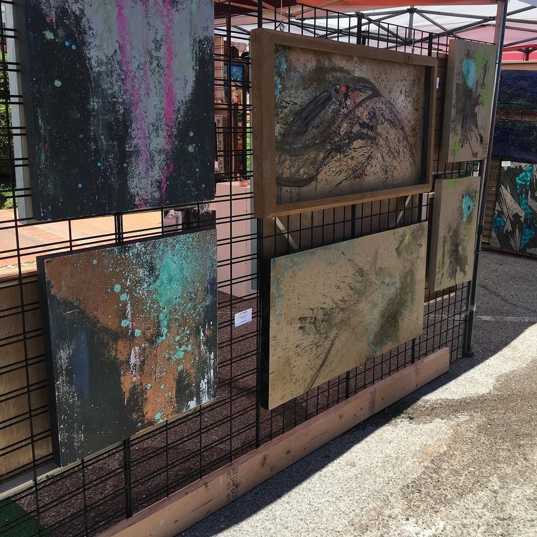 Abstract art paintings displayed on a metal grid, outdoors. Earth tones, textures, and splashes of color.