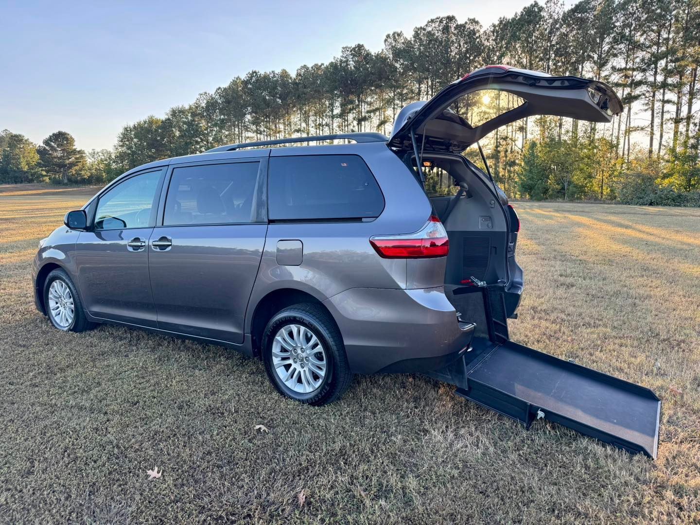 Gray wheelchair-accessible minivan with ramp extended, parked on grass.