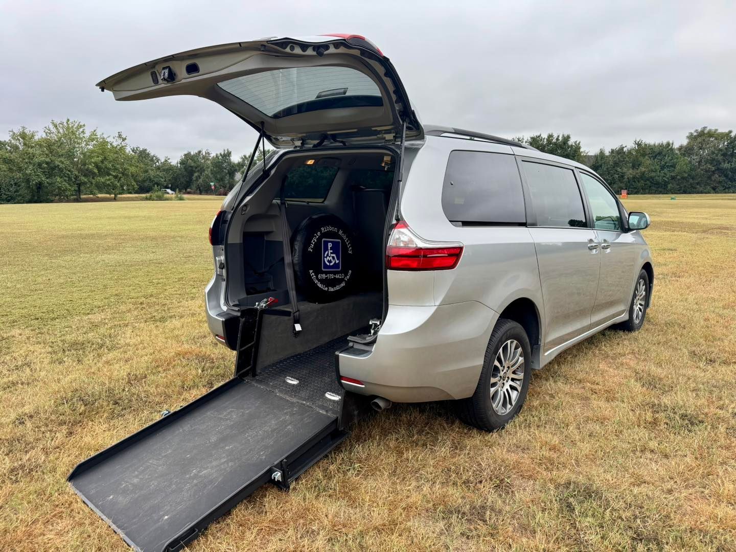 Silver wheelchair-accessible van with open ramp on grassy field.
