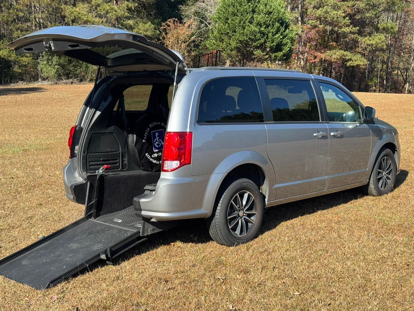 Gray minivan with a wheelchair ramp extended on a grassy field, rear door open.