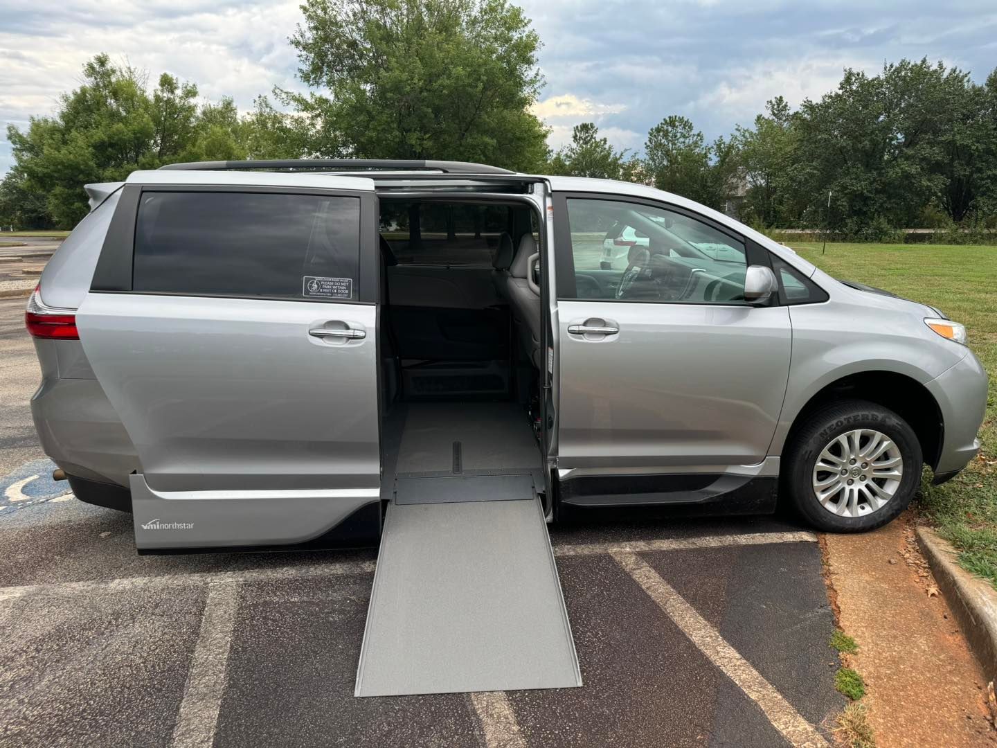 Silver wheelchair-accessible van with side ramp extended, parked in a lot. The door is open.