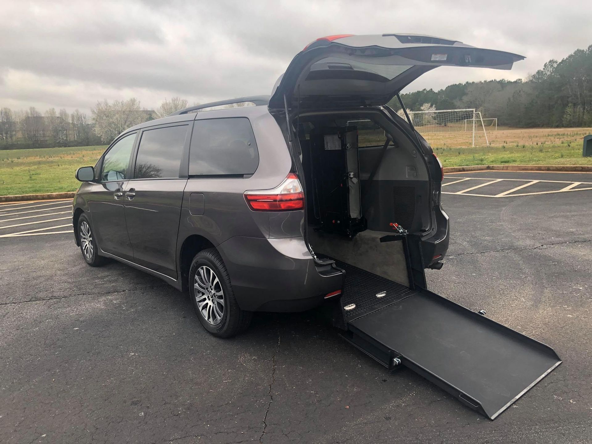 Dark gray minivan with ramp deployed, parked on asphalt, cloudy day.
