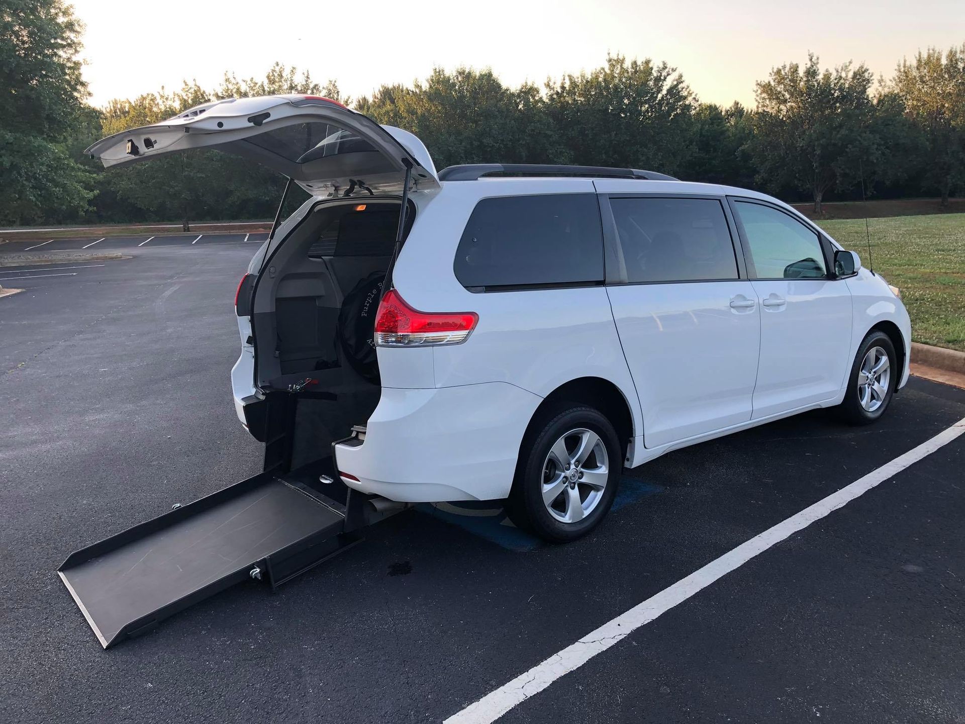 White minivan with a ramp deployed in a parking lot. The rear door is open.