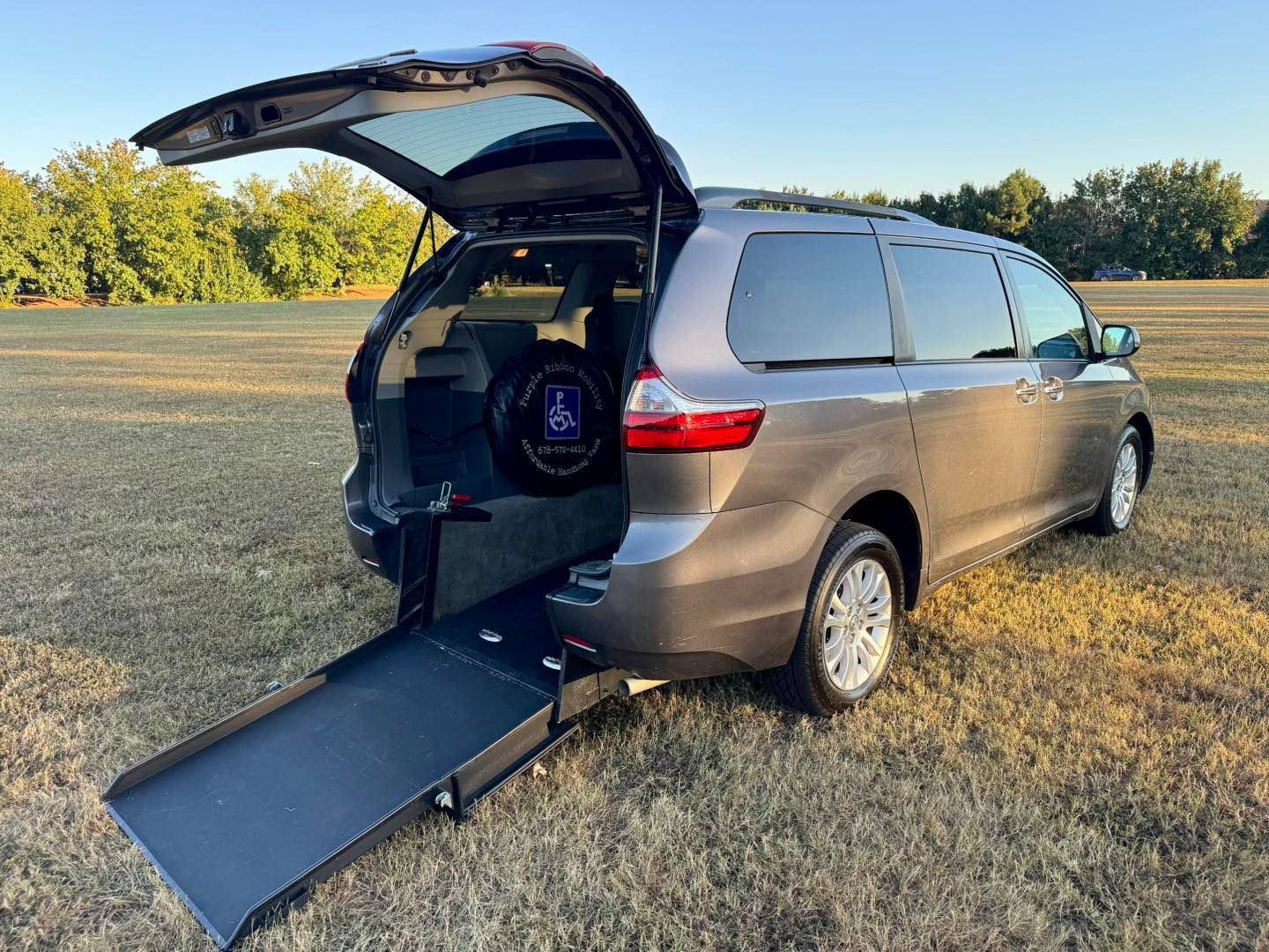 Gray minivan with wheelchair ramp deployed, parked on grass. Rear door open.