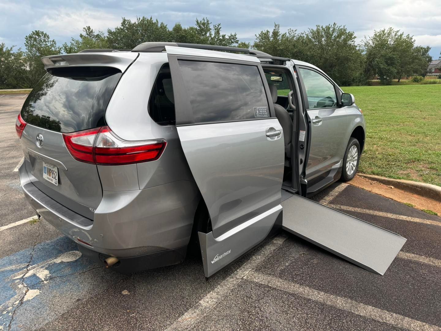 Silver accessible van with ramp extended, parked on asphalt, cloudy sky in background.