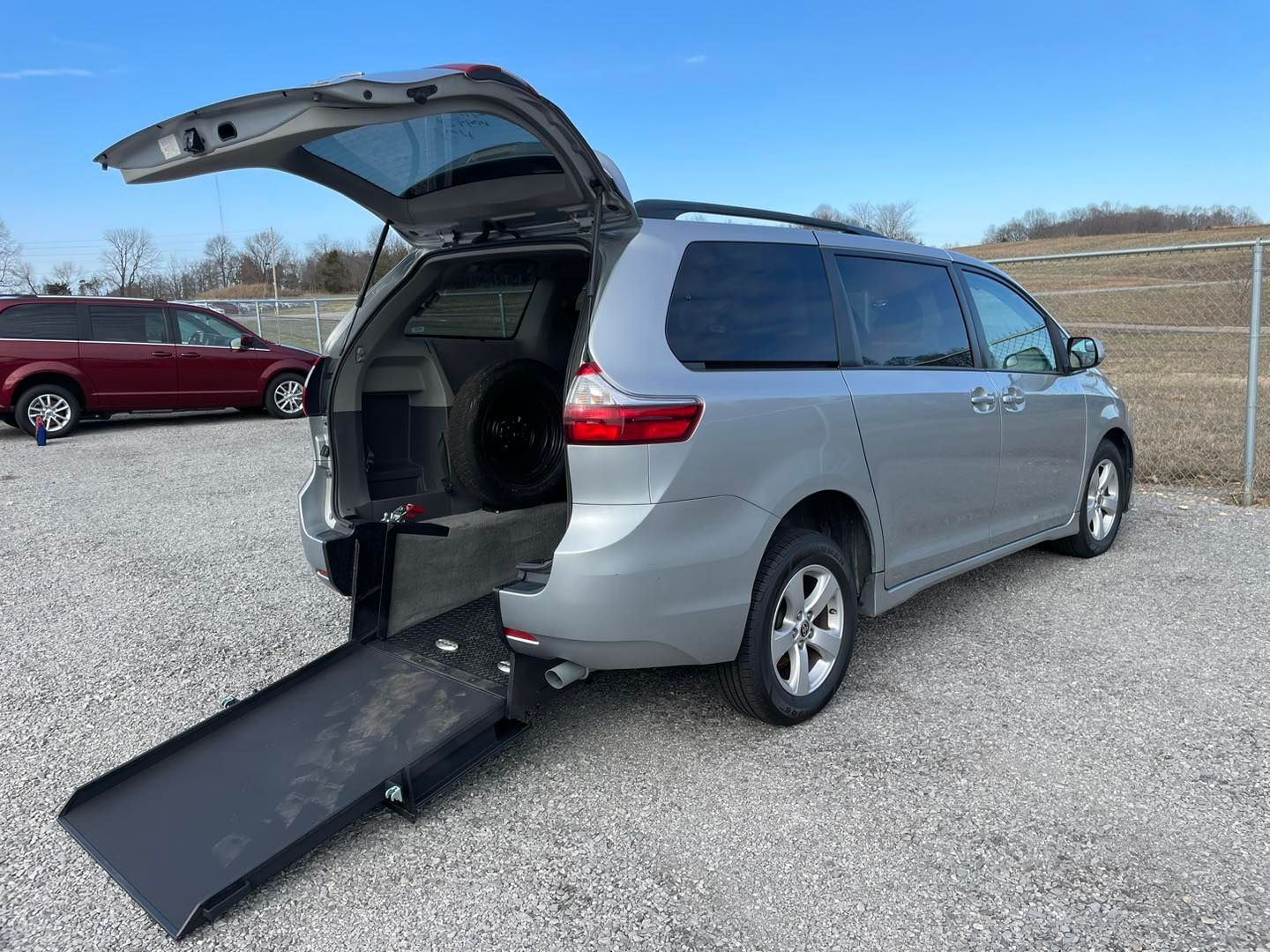 Silver wheelchair-accessible minivan with ramp extended. Parked outdoors, ramp lowered, rear hatch open.