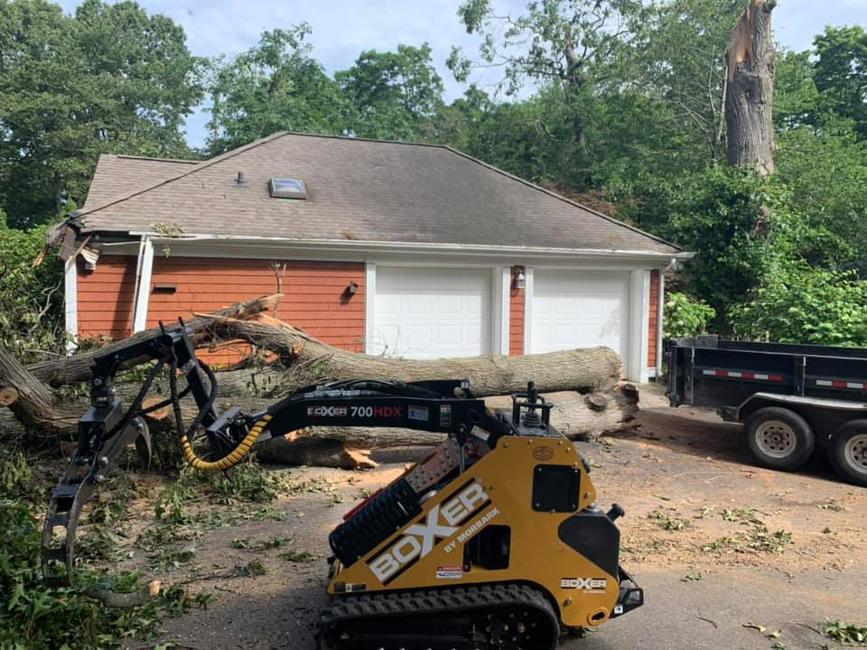 Boxer machine removing a fallen tree from a garage with orange siding, trailer in the background.