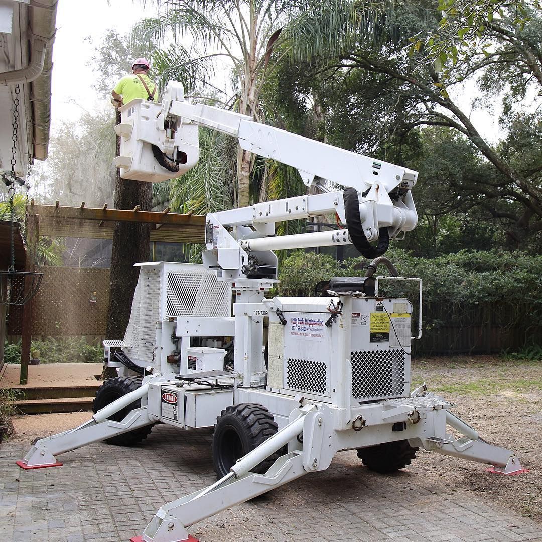 Man in bucket lift trimming trees near a building, lift is white, in an outdoor setting.