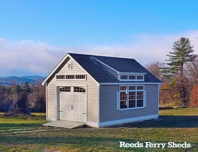A beige shed with a garage door, side windows, and a dormer roof, situated in a field under a clear blue sky.