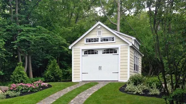 A small, light yellow detached garage with a white door and windows, surrounded by green trees and landscaped gardens.