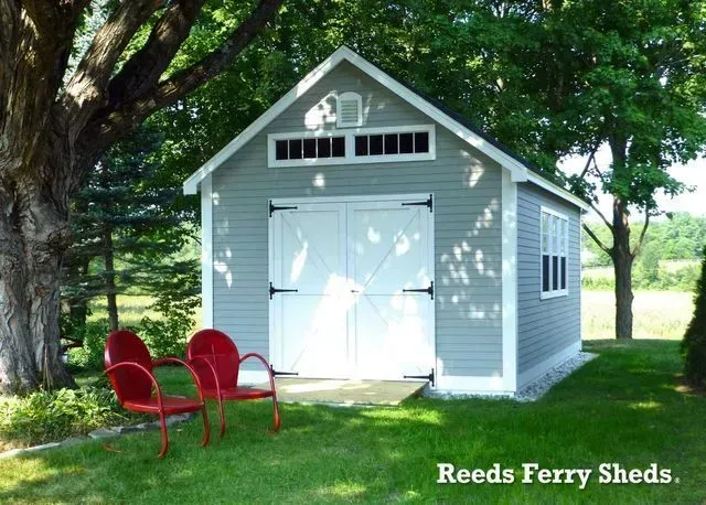 A grey shed with white trim and double doors sits in a grassy yard, with two red chairs in the foreground.