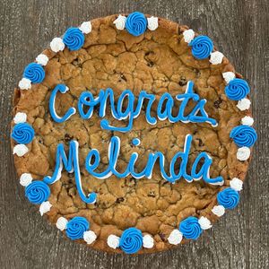 Round cookie with blue and white frosting reading “Congrats Melinda” on a wooden surface