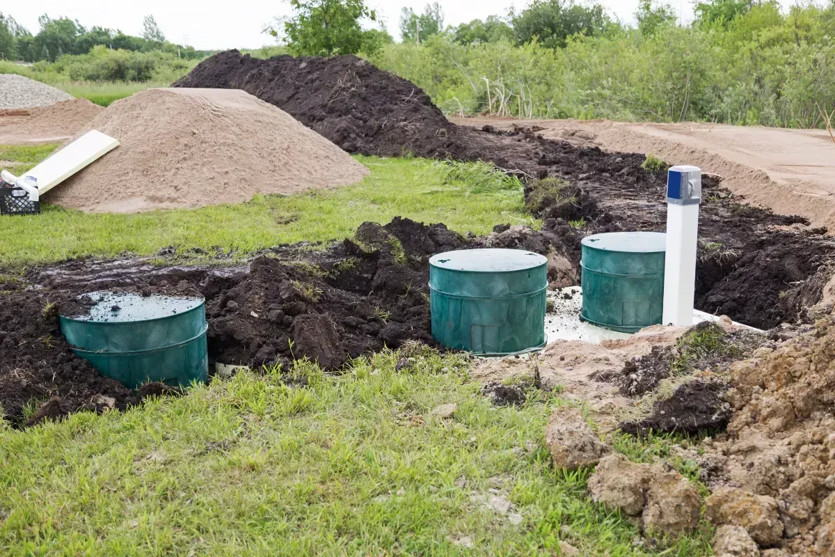 Three green septic tanks in a dug trench with dirt and sand piles.