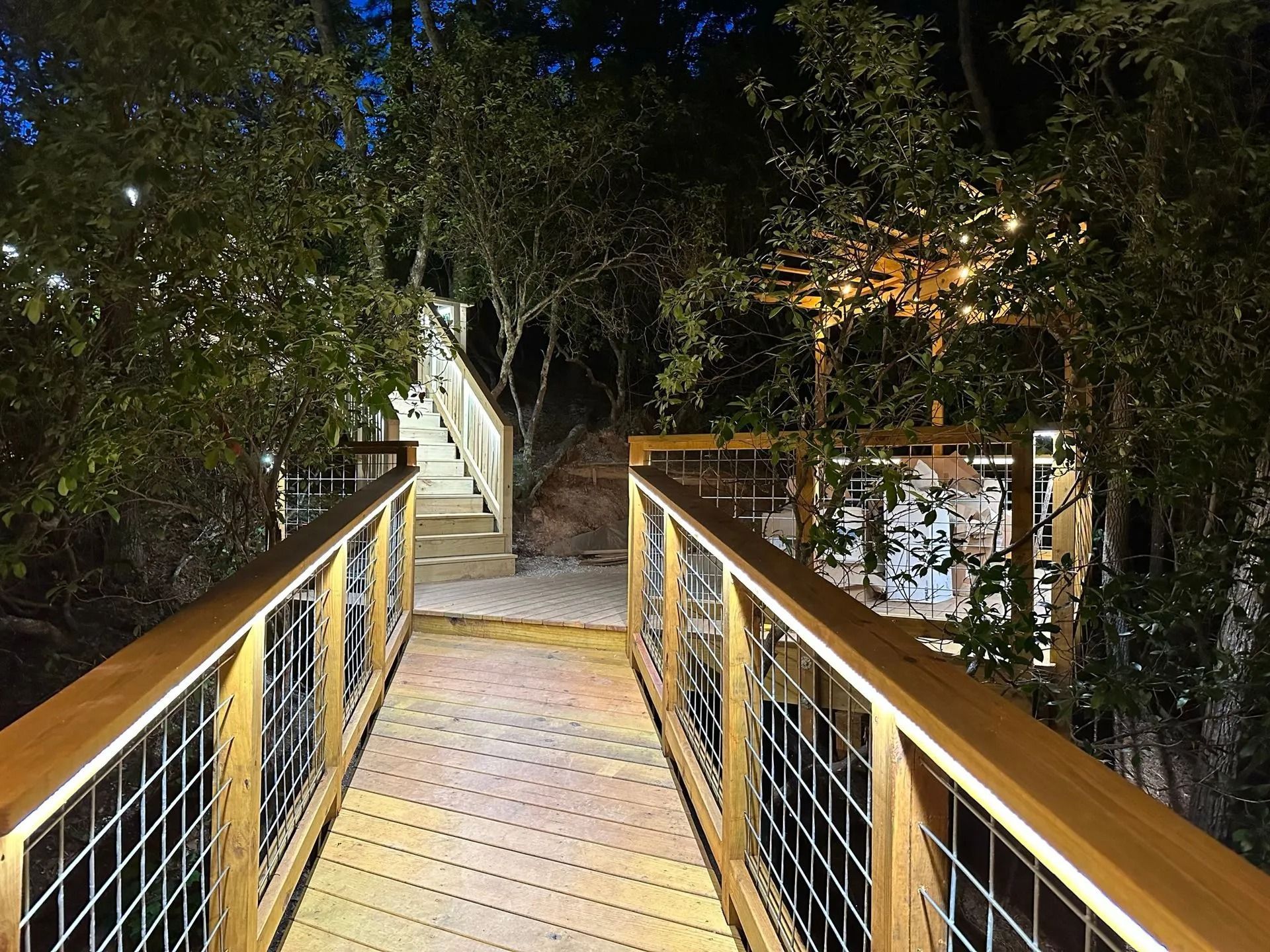 Wooden elevated walkway with metal railings lit at night, leading to an open-air structure.