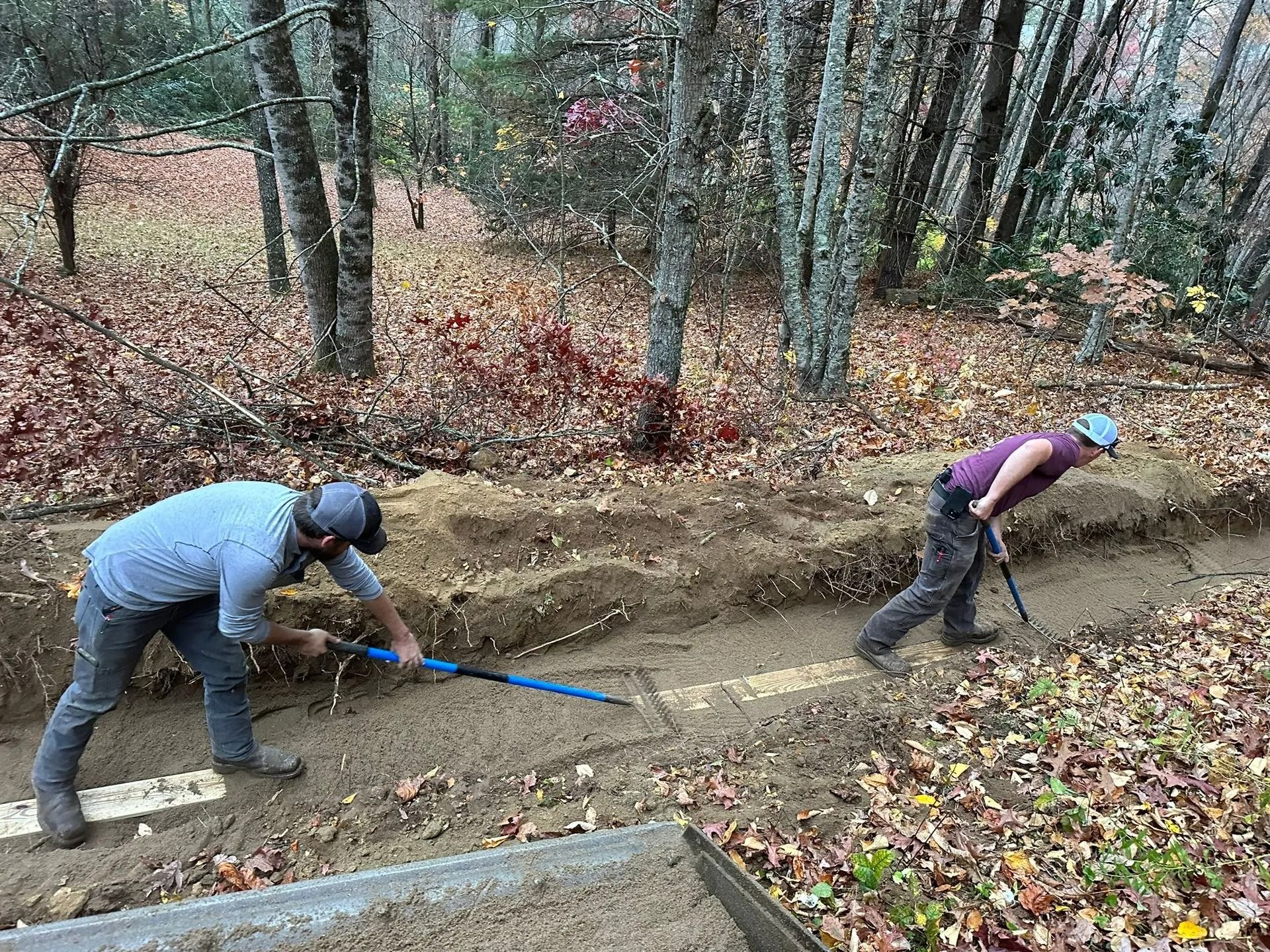 Two people digging a trench in a wooded area, likely for utility pipes.