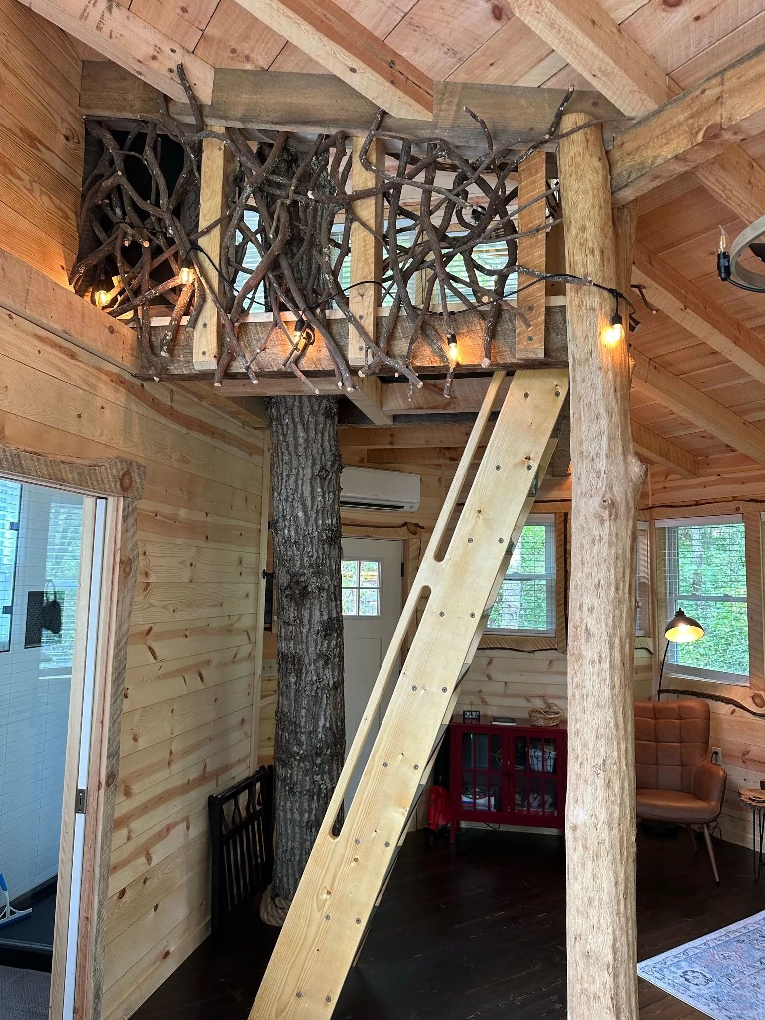 Interior of a rustic treehouse with a loft, wooden ladder, and natural wood beams.
