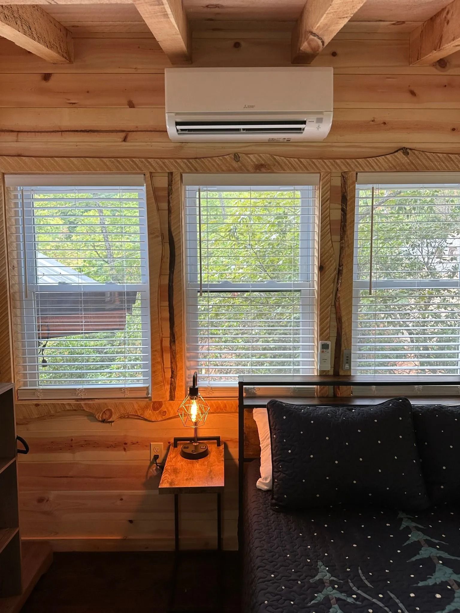 Bedroom interior with wooden walls, windows with blinds, air conditioner, bed, and nightstand with lamp.